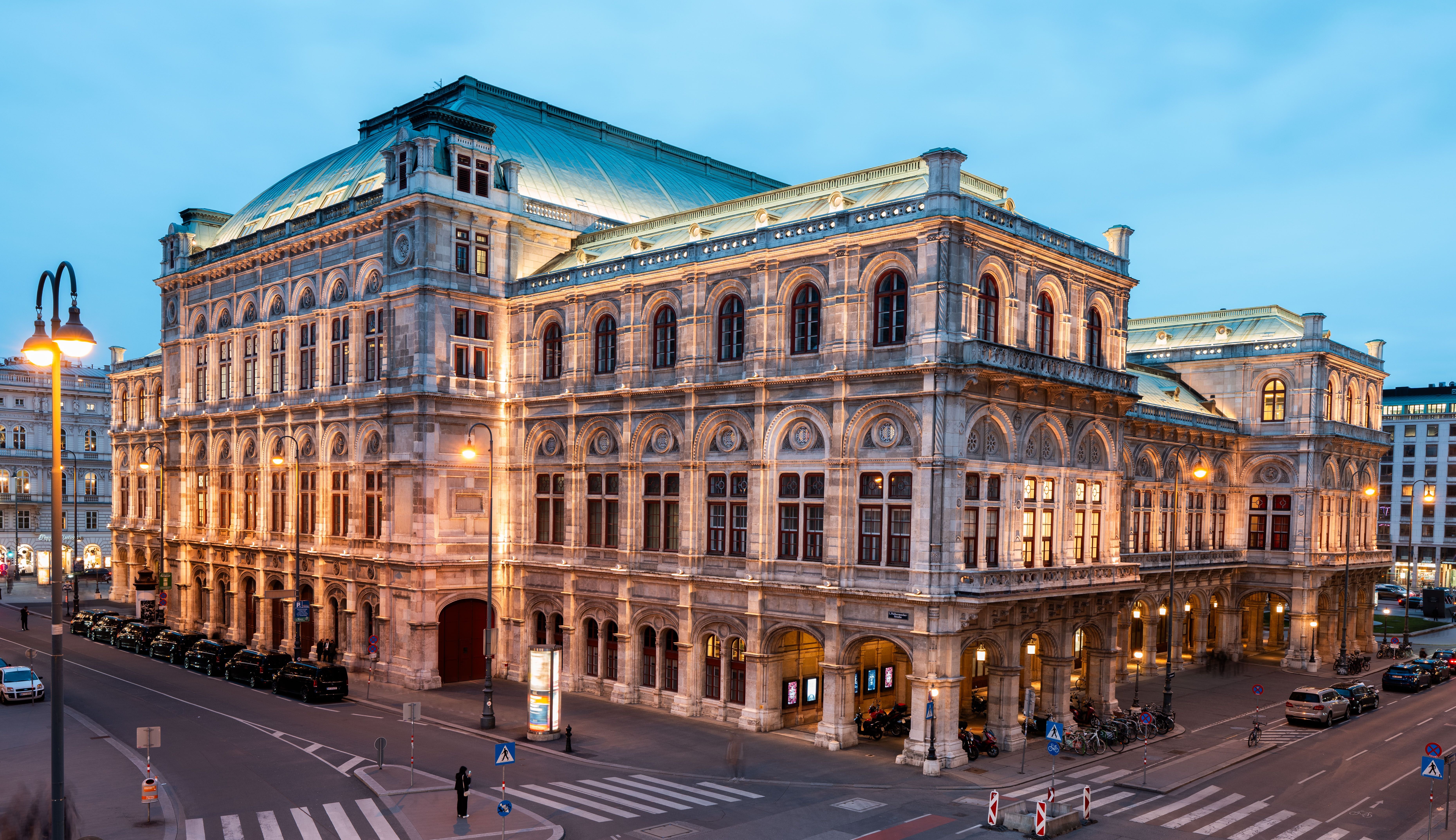 Die Wiener Staatsoper bei Abenddämmerung, außen angestrahlt und von Straßen umgeben.
