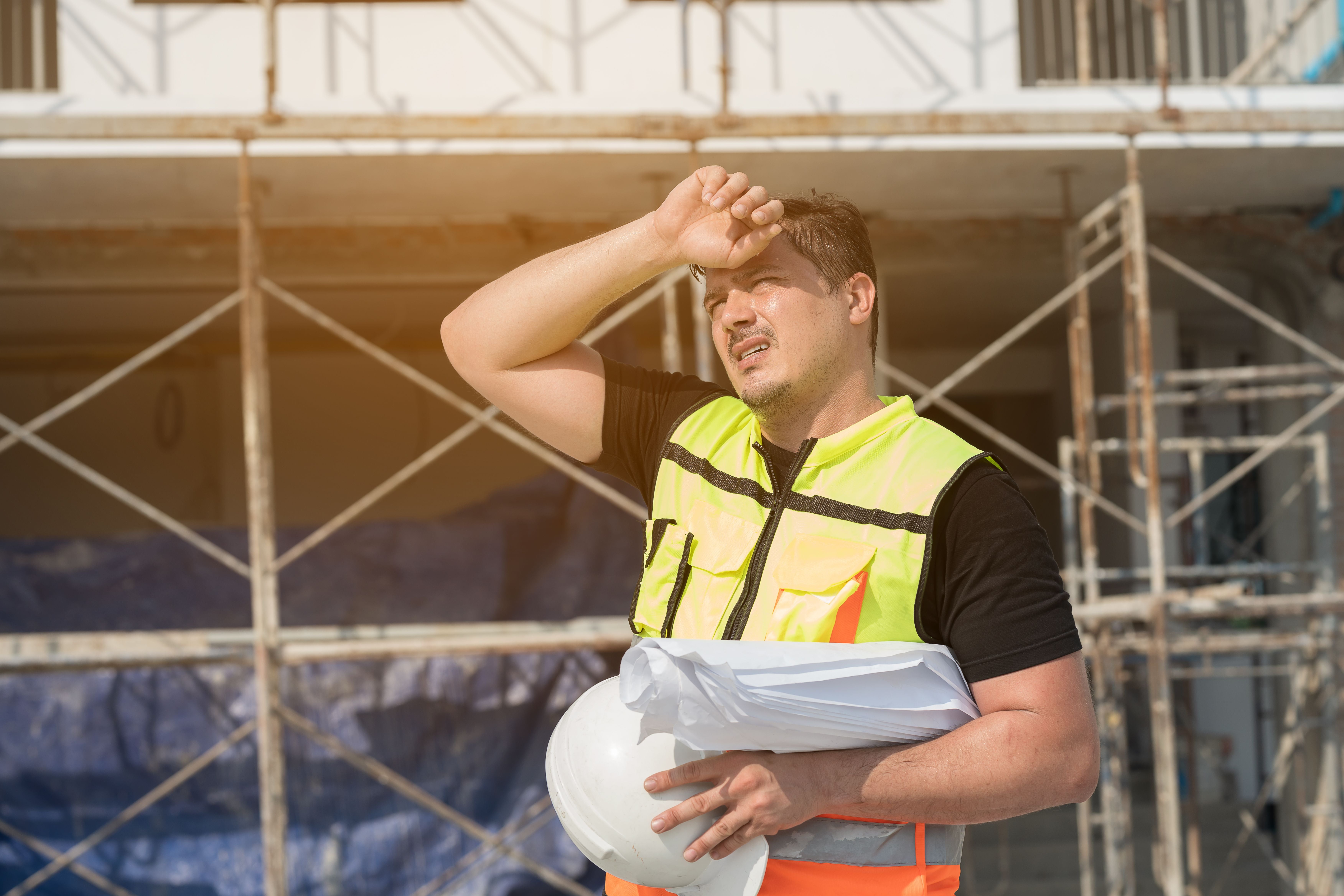 Müder Bauarbeiter im Warnwesten wischt sich auf der Baustelle den Schweiß von der Stirn.