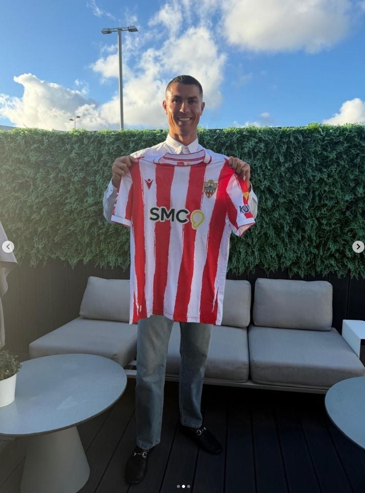 Ein Mann hält ein rot-weiß gestreiftes Fußballtrikot und lächelt in die Kamera.