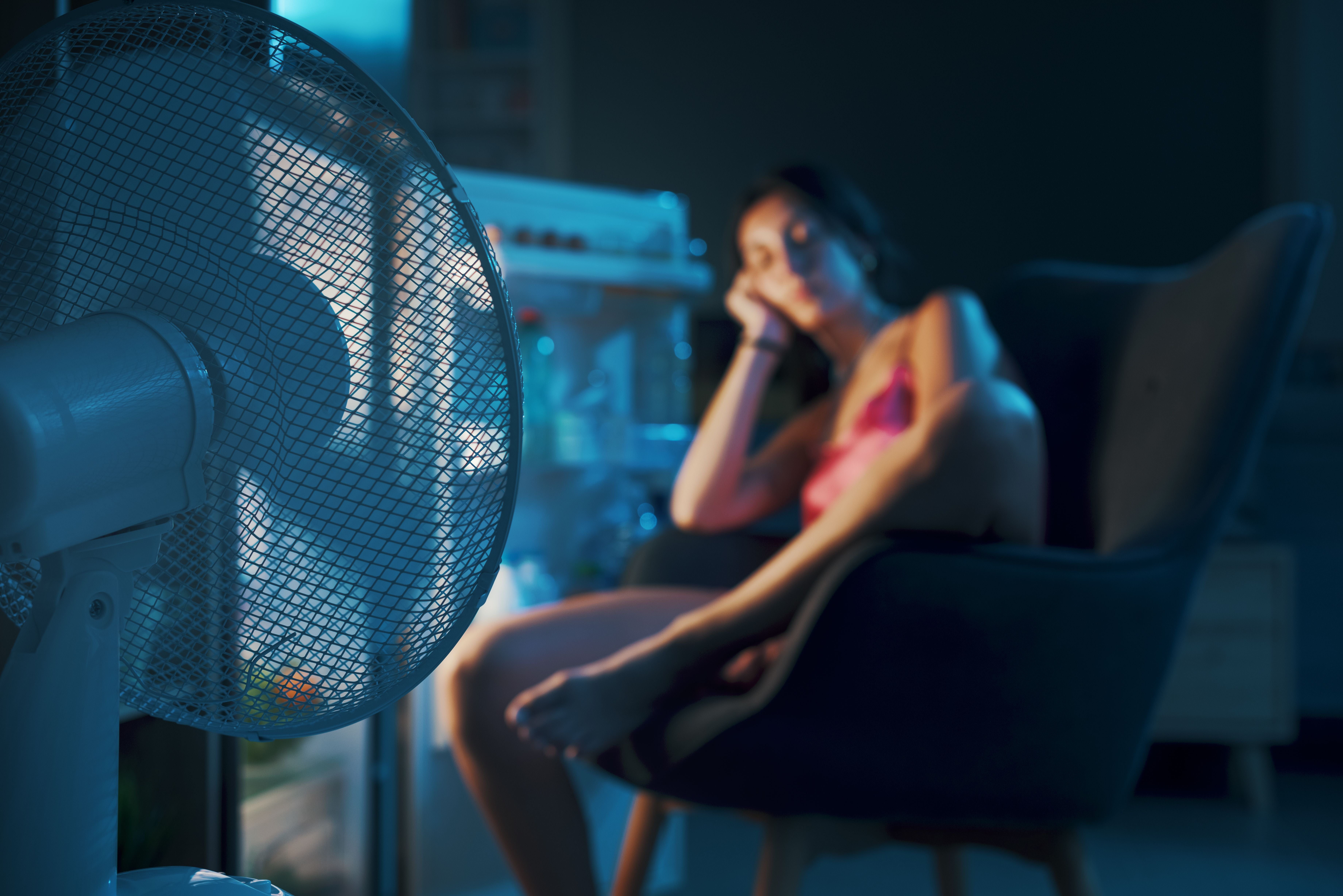 Erschöpfte Frau kühlt sich vor offenem Kühlschrank mit Ventilator während Sommerhitze ab.