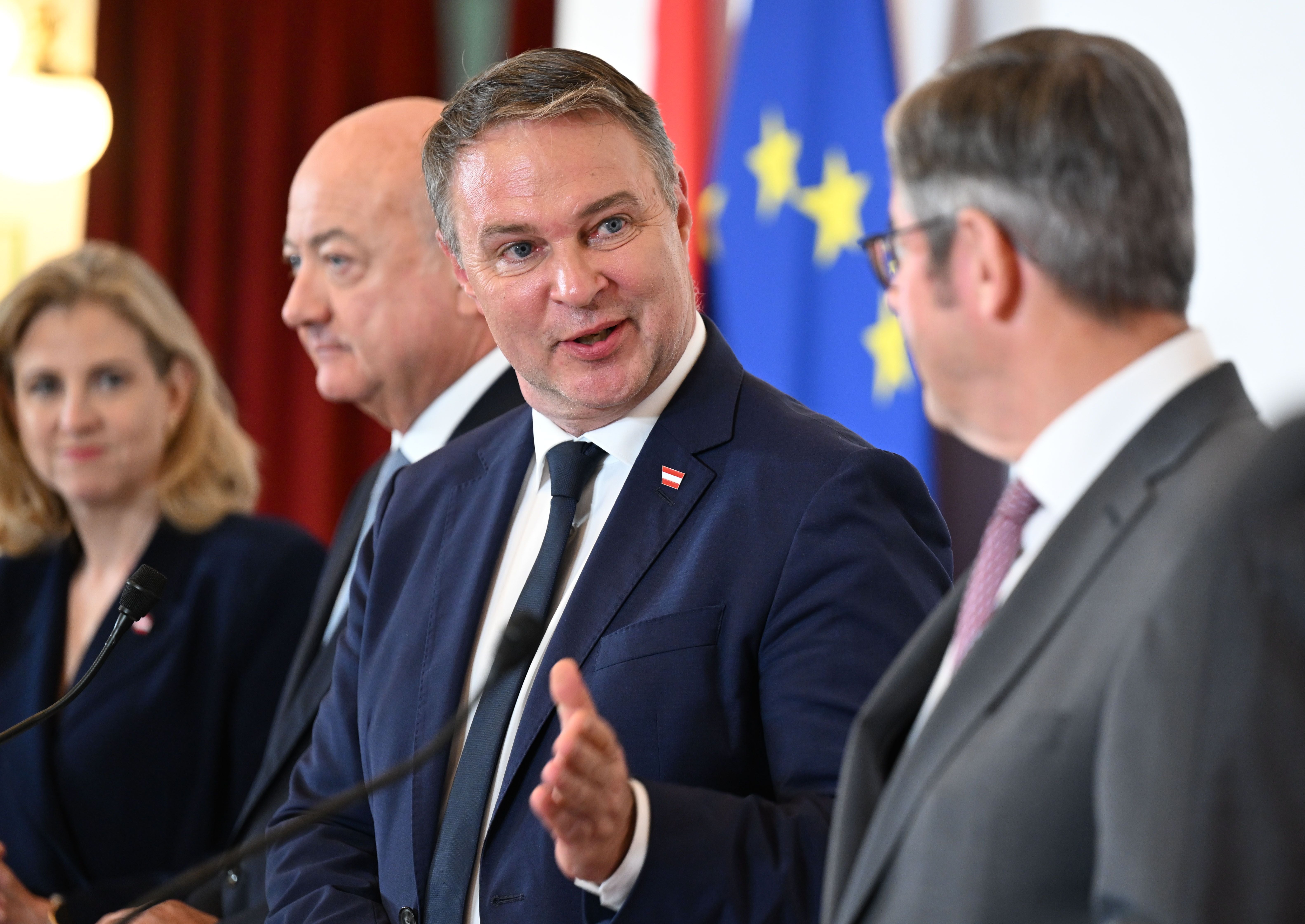 Vier Personen im Anzug sprechen bei einer Pressekonferenz vor EU- und Österreich-Flaggen.