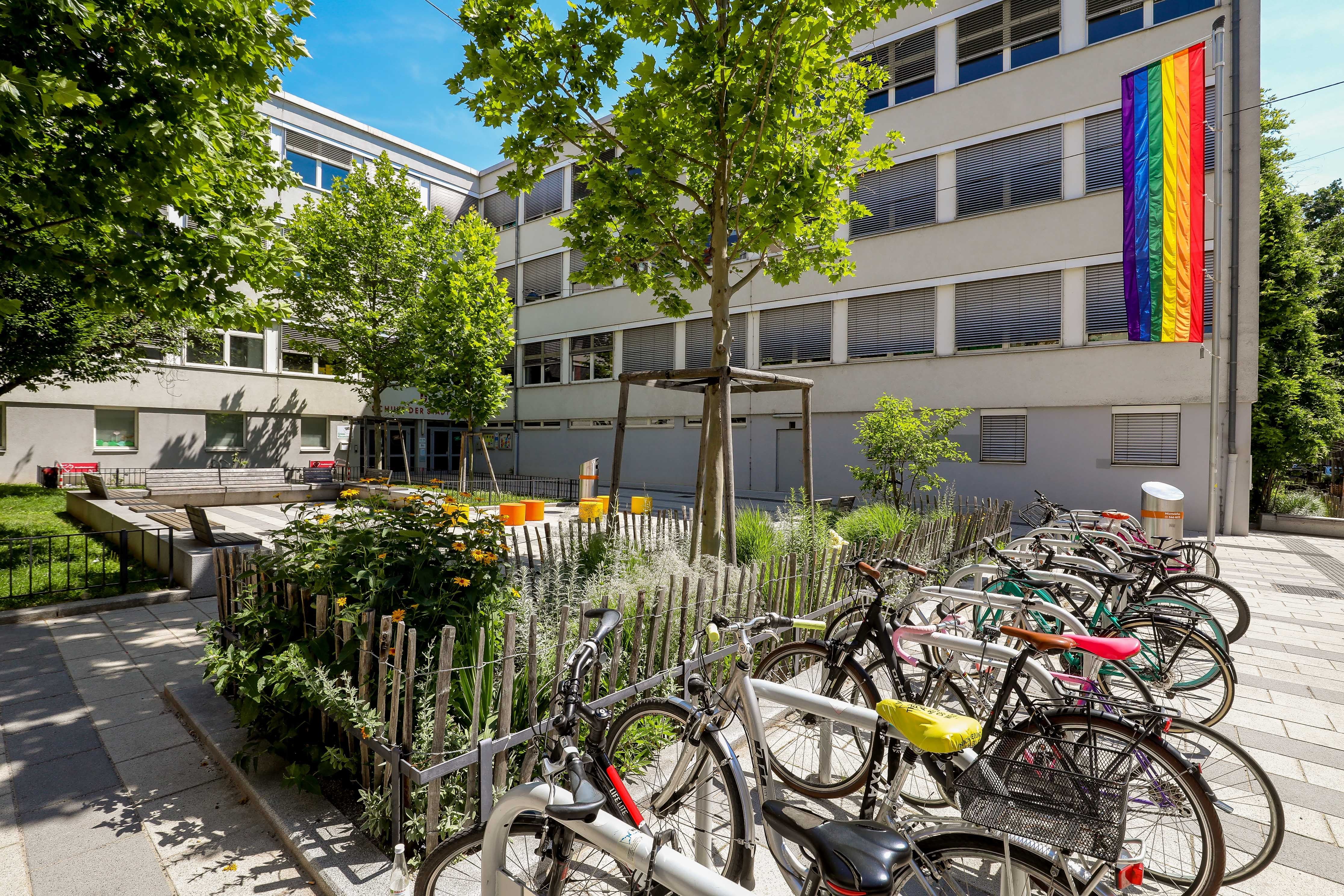 Fahrradständer mit Fahrrädern vor einer Schule mit Regenbogenflagge und viel Grün.