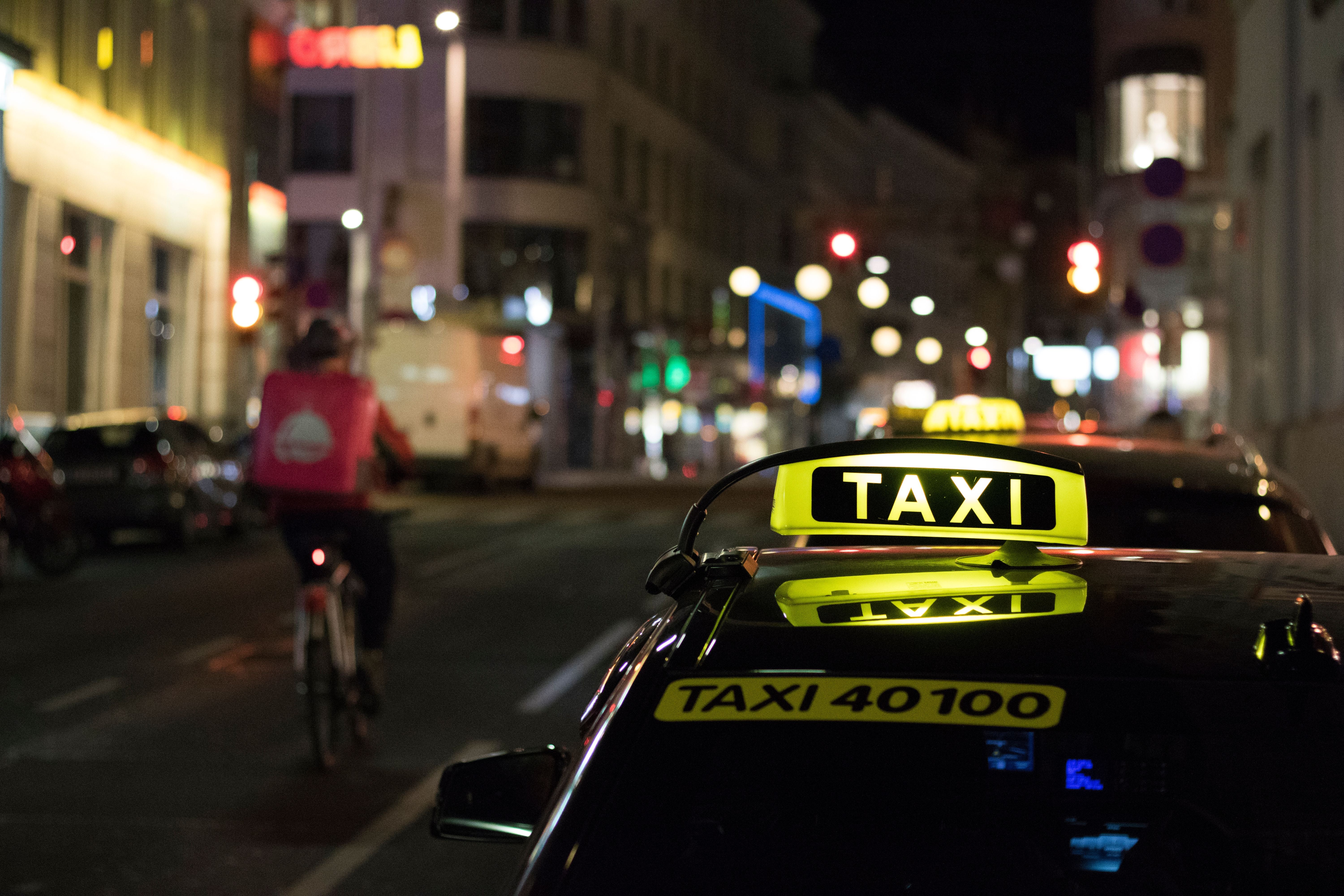 Ein beleuchtetes Taxi steht nachts auf einer Straße, vorbeifahrender Kurier auf Fahrrad.