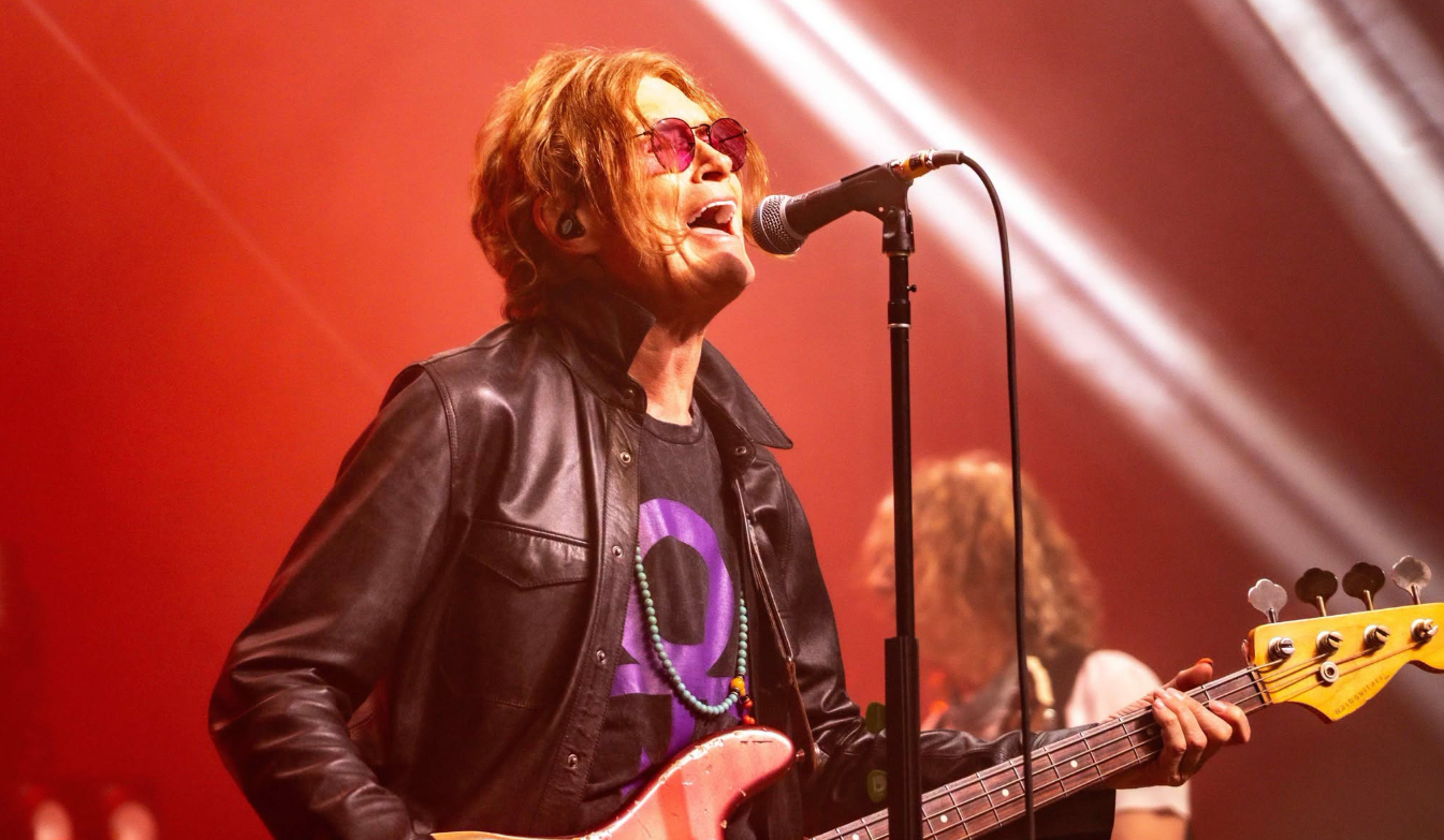 Sänger mit Sonnenbrille und Lederjacke singt auf der Bühne und spielt Bassgitarre.