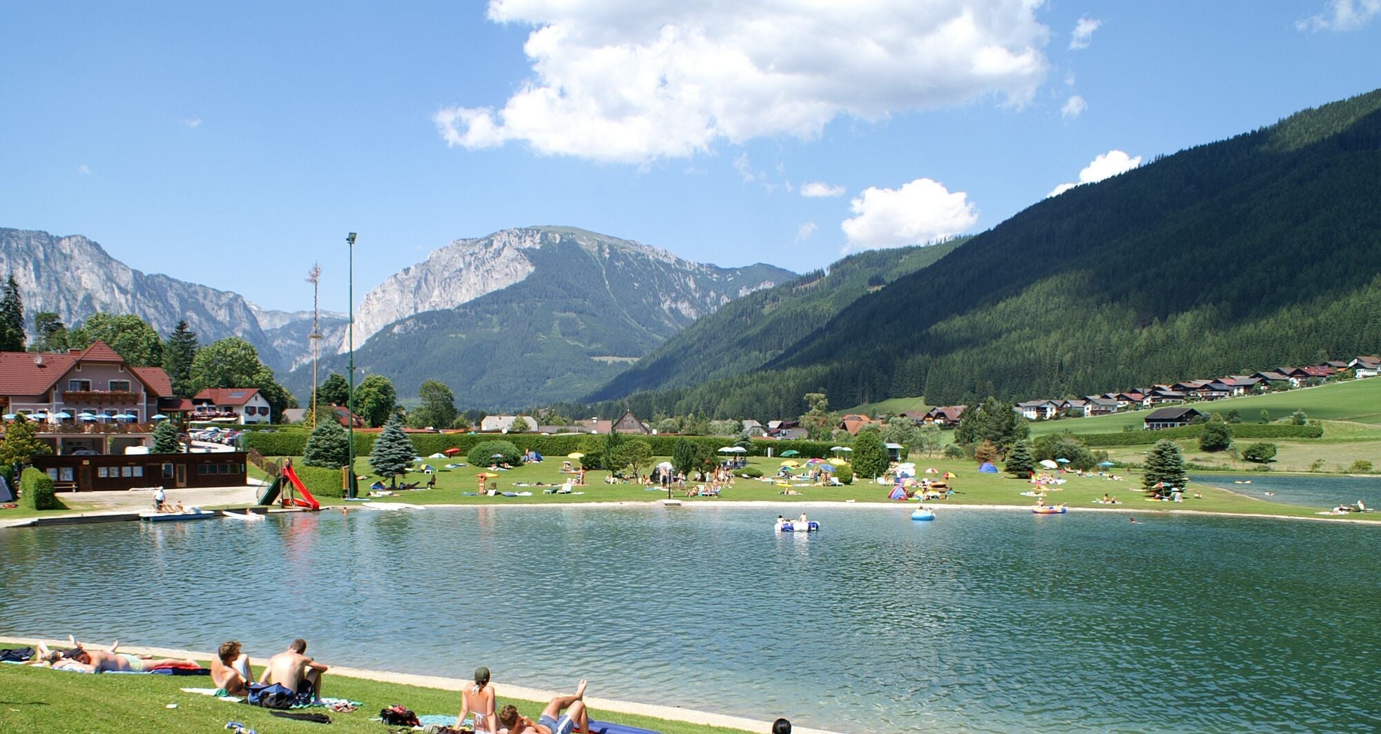 Menschen entspannen am Badesee mit Wiese, umliegenden Häusern und Bergen im Hintergrund.