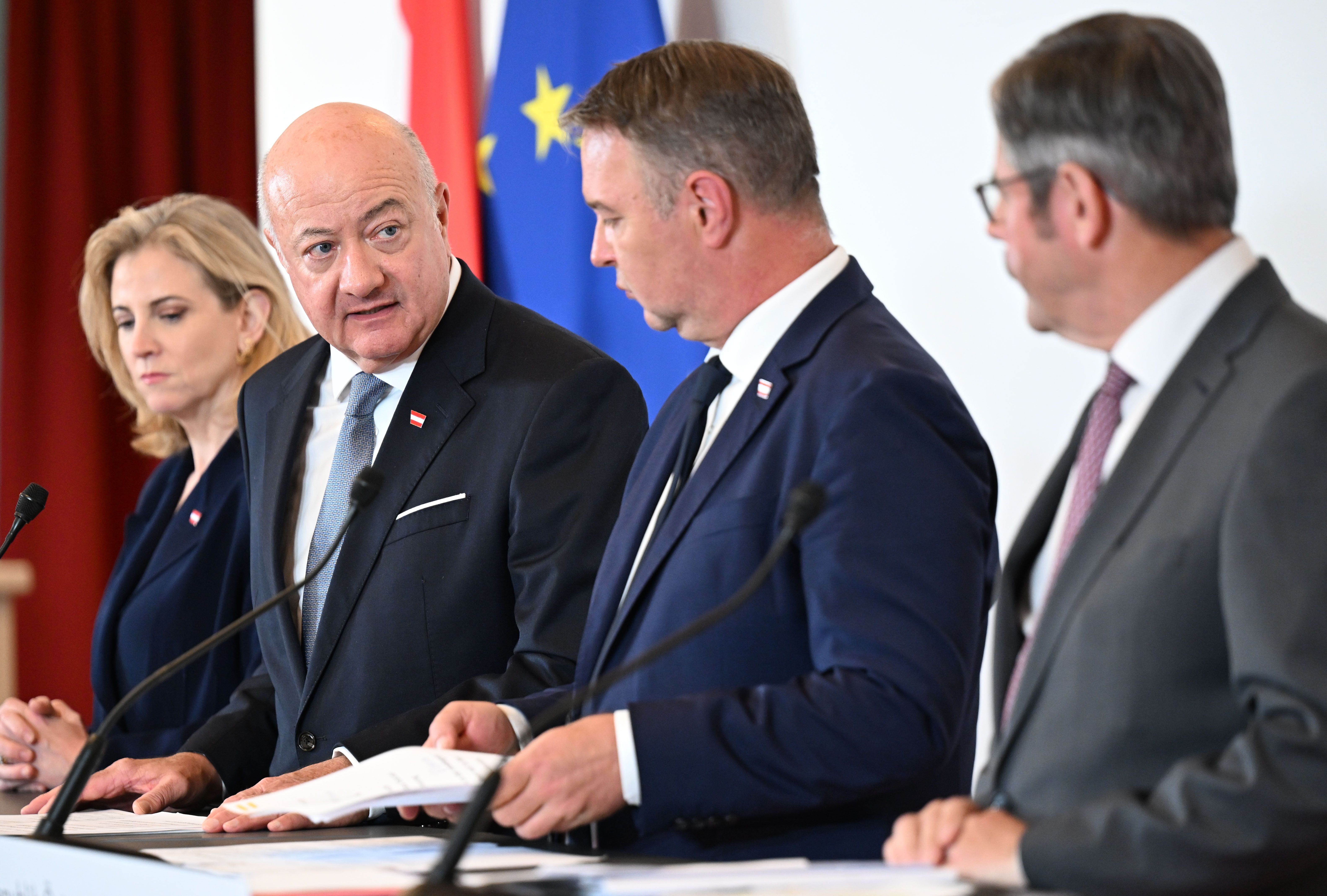 Vier Personen stehen bei einer Pressekonferenz vor Mikrofonen und sprechen miteinander.
