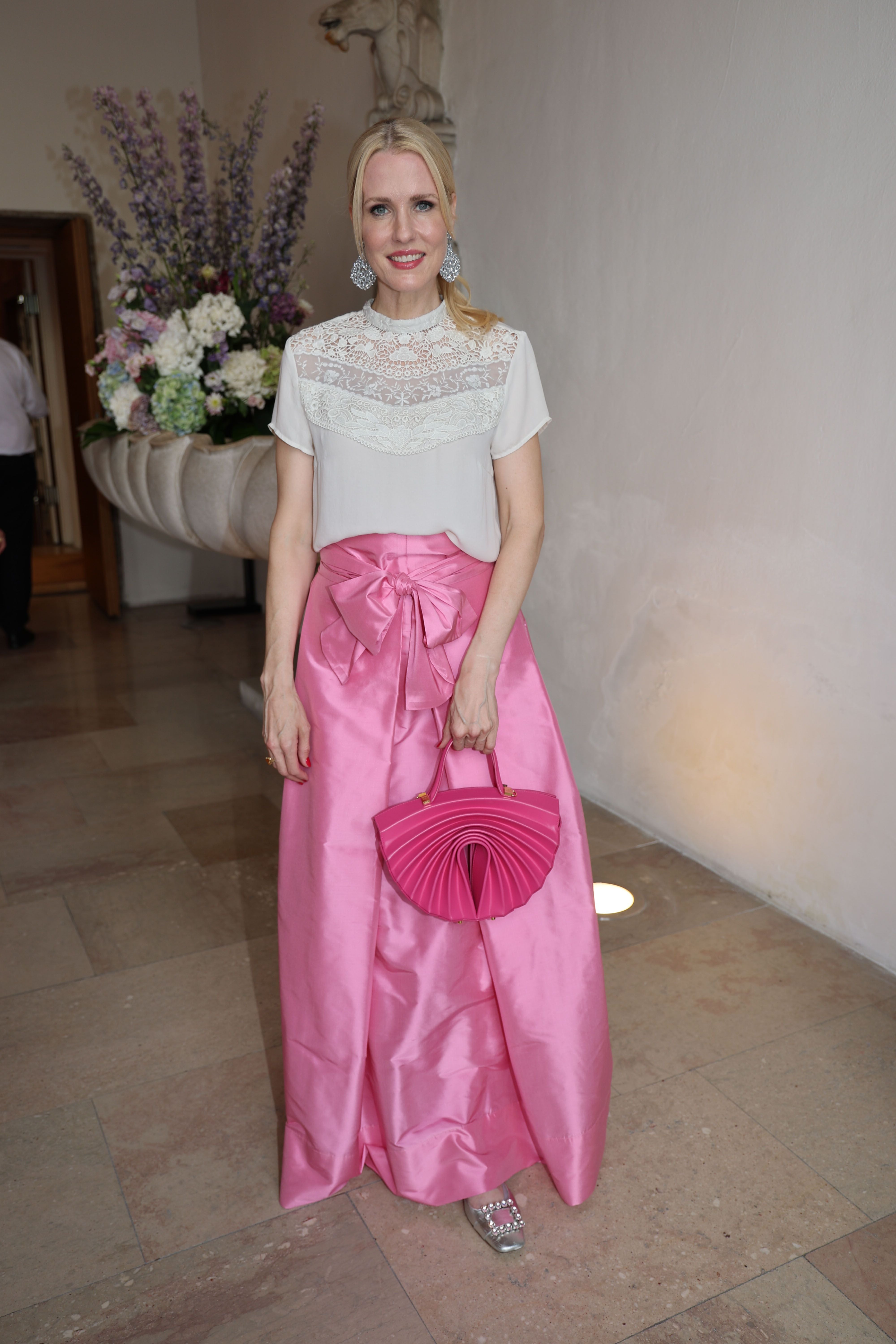 Frau in weißer Spitzenbluse und rosa Abendrock mit passender pinker Handtasche, stehend im Foyer.