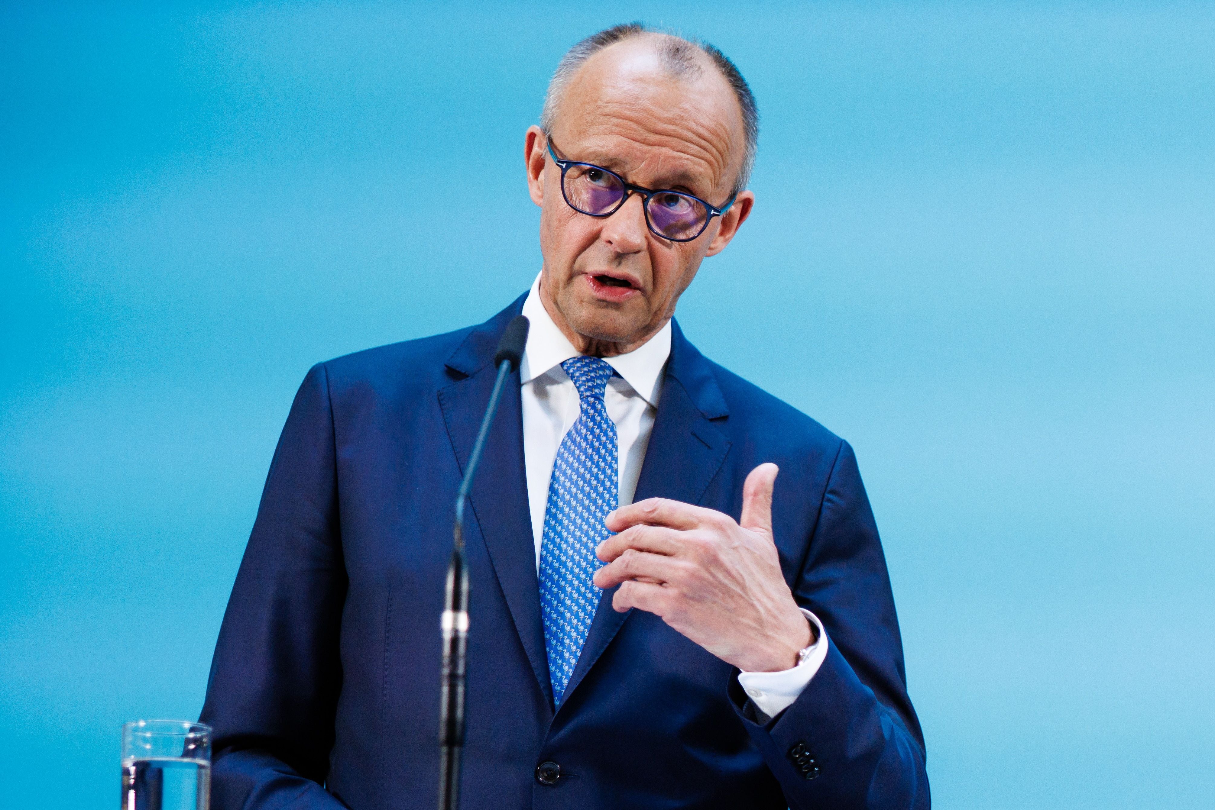Ein Mann im Anzug spricht bei einer Pressekonferenz vor blauem Hintergrund.