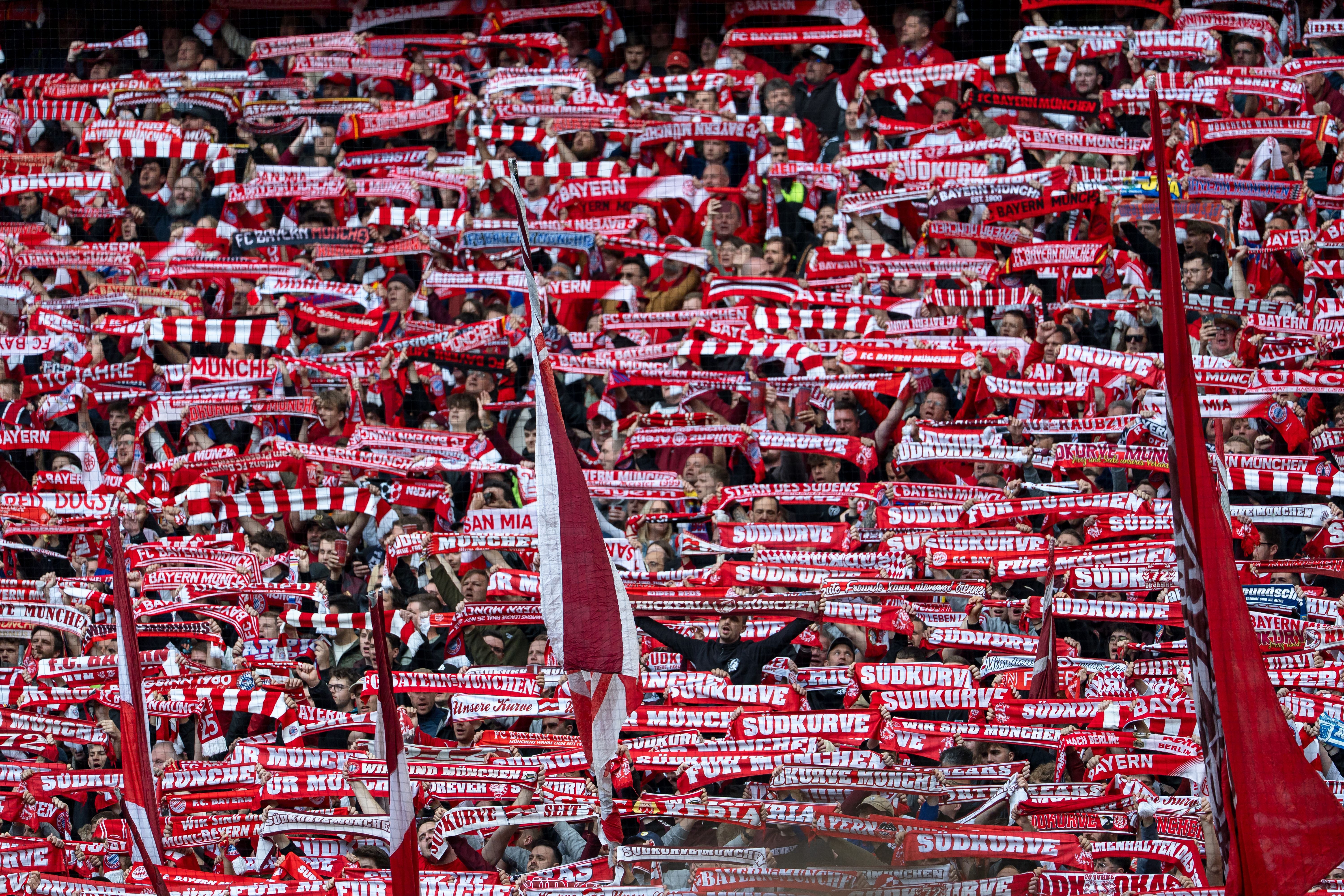 Fans von Bayern München halten rote und weiße Schals in einer vollen Stadionkurve hoch.