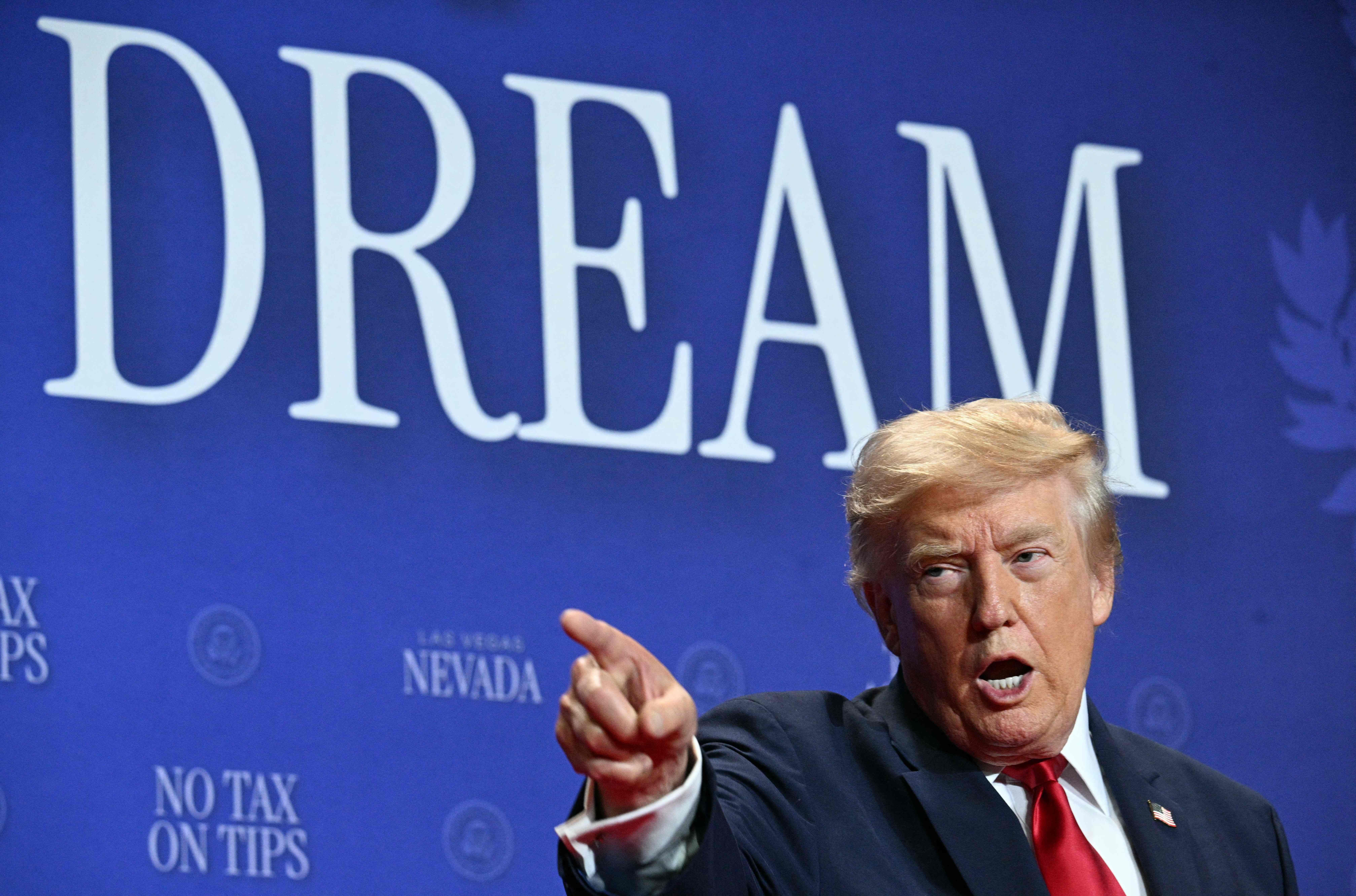 Ein Mann im Anzug spricht und gestikuliert vor dem Wort "DREAM" auf blauem Hintergrund.