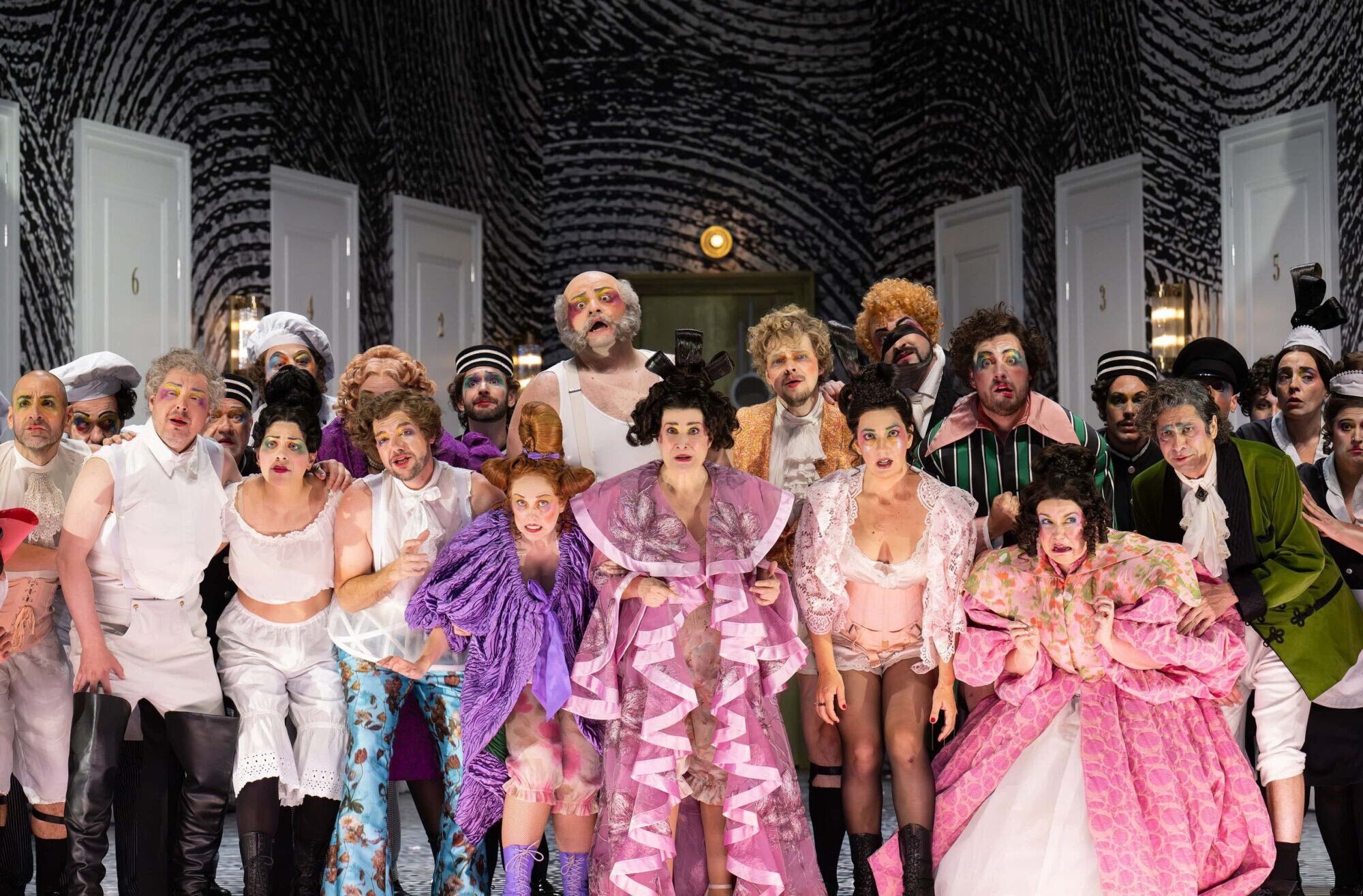 Ensemble in bunten, barocken Kostümen und buntem Make-up auf einer Theaterbühne.
