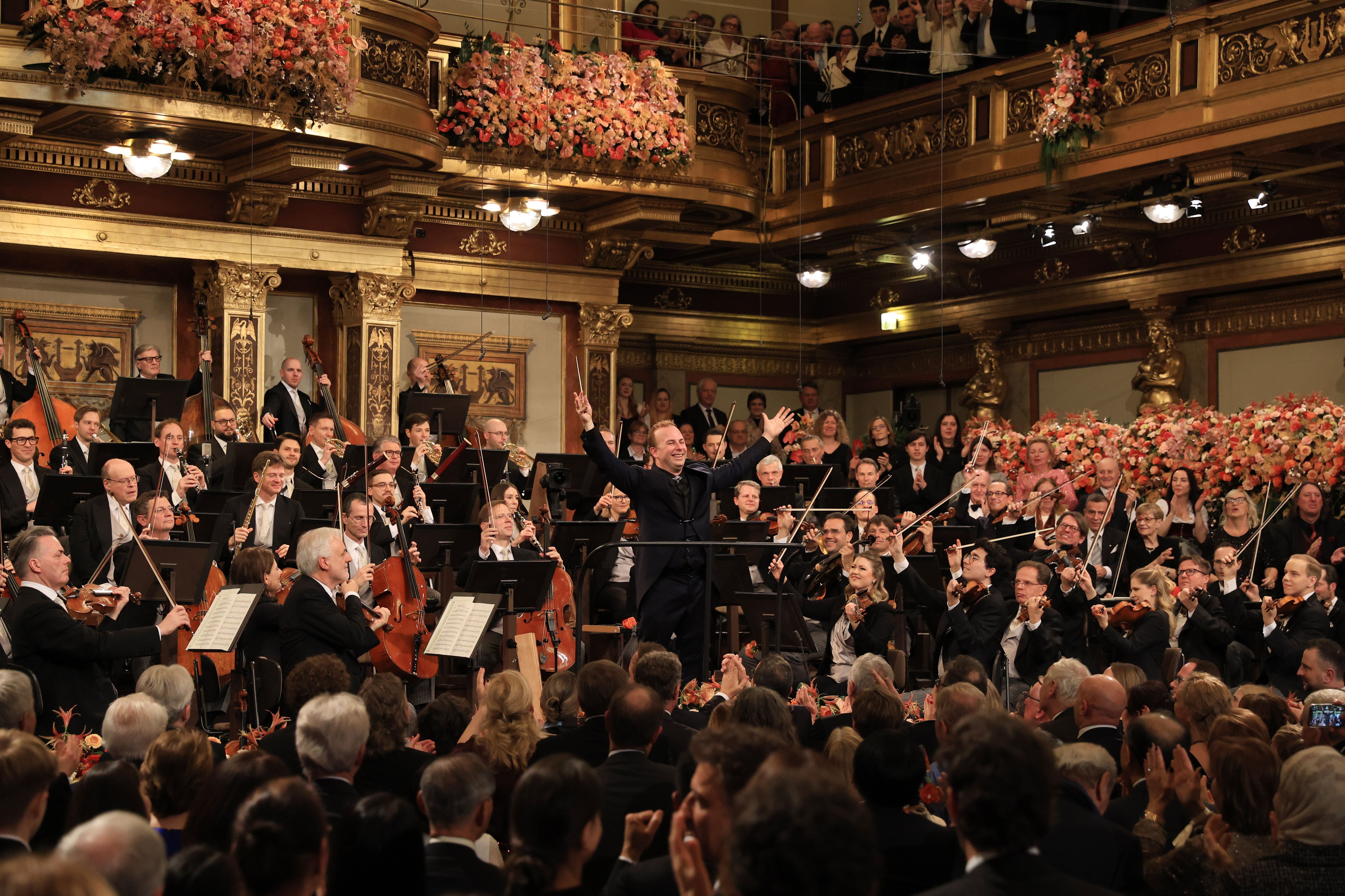 Orchester und Dirigent im goldenen Konzertsaal, Publikum applaudiert, Blumendekoration im Hintergrund.