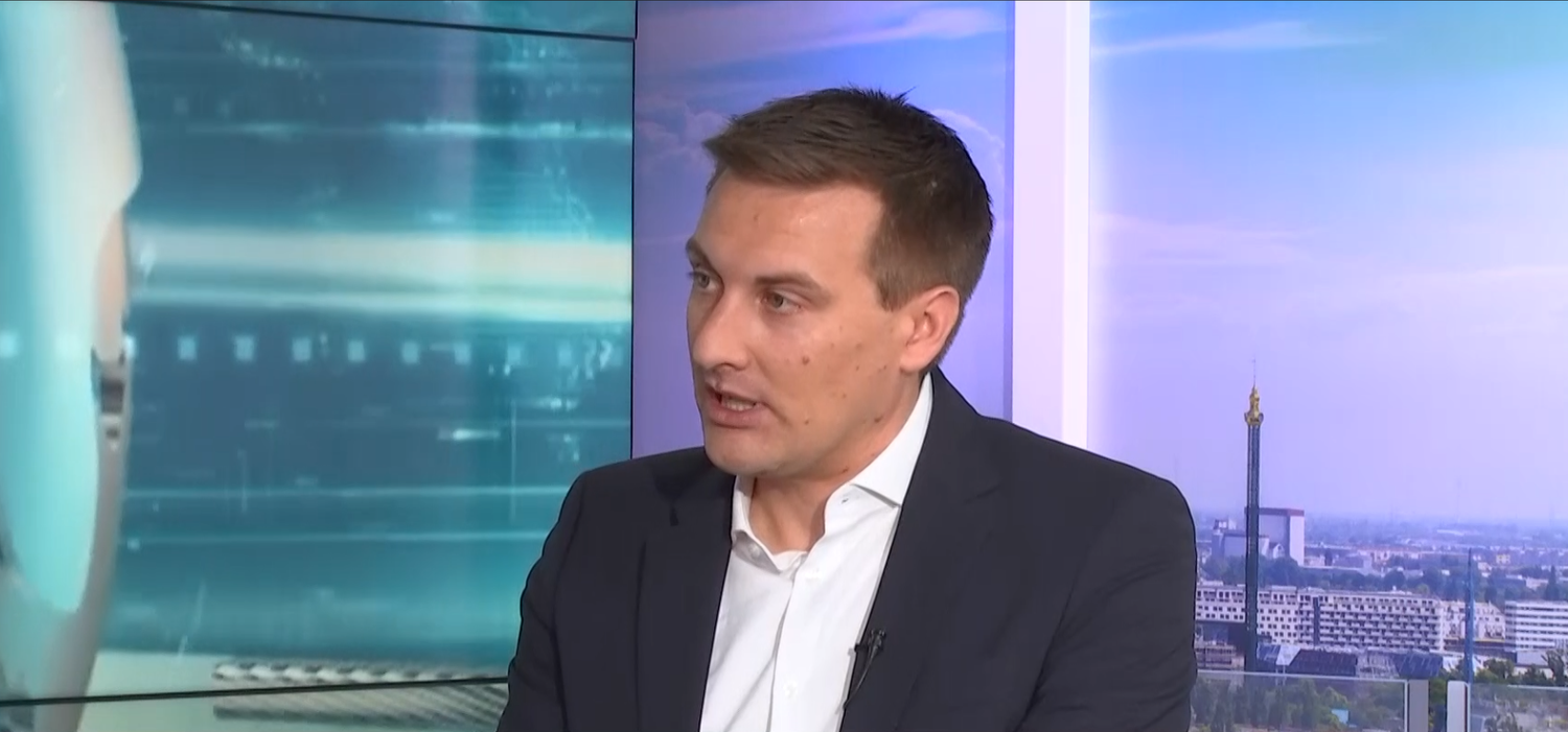 Ein Mann im Anzug spricht in einem Fernsehstudio, Stadtpanorama im Hintergrund.