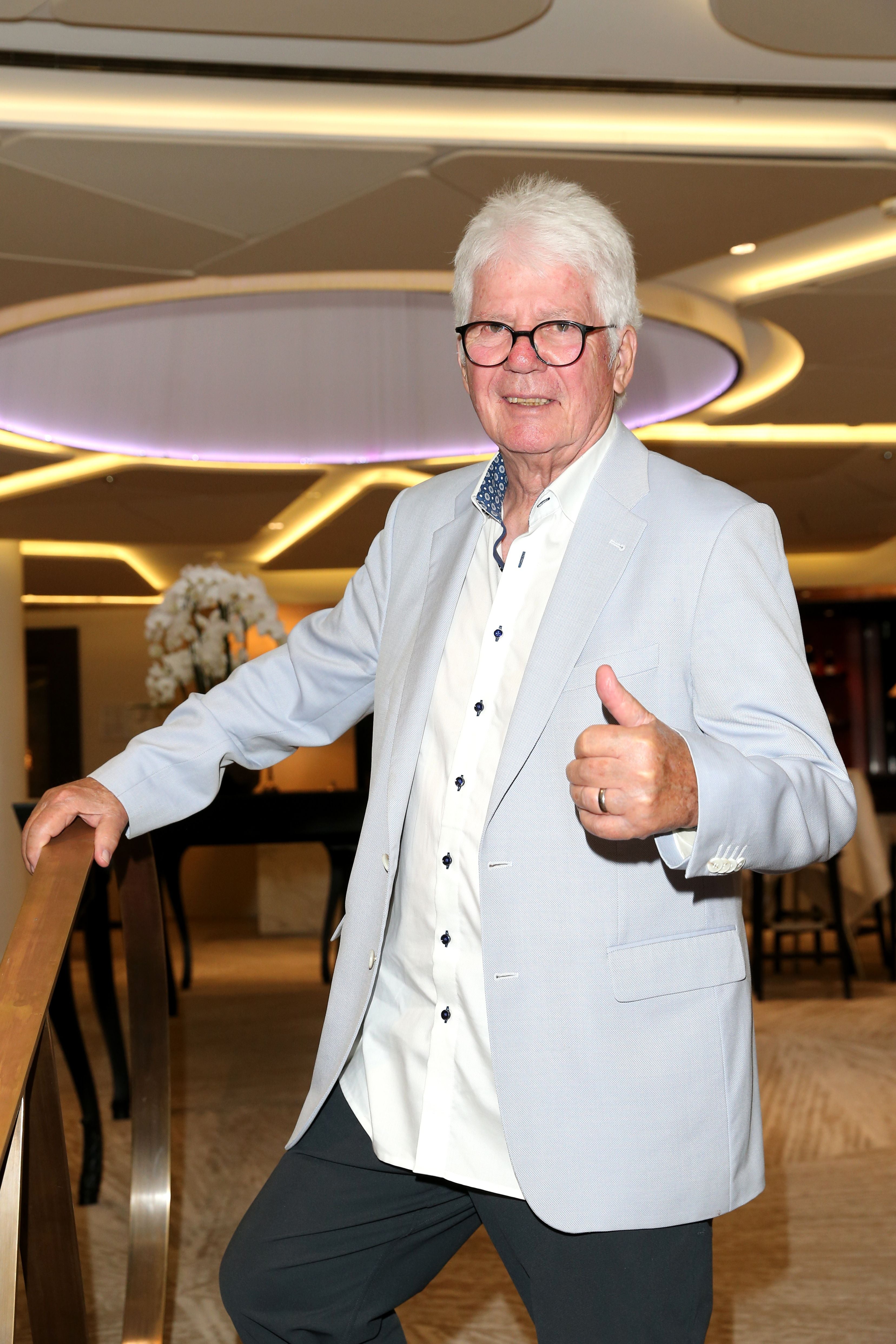 Älterer Mann mit Brille zeigt Daumen hoch und lächelt, im eleganten Innenraum eines Hotels.