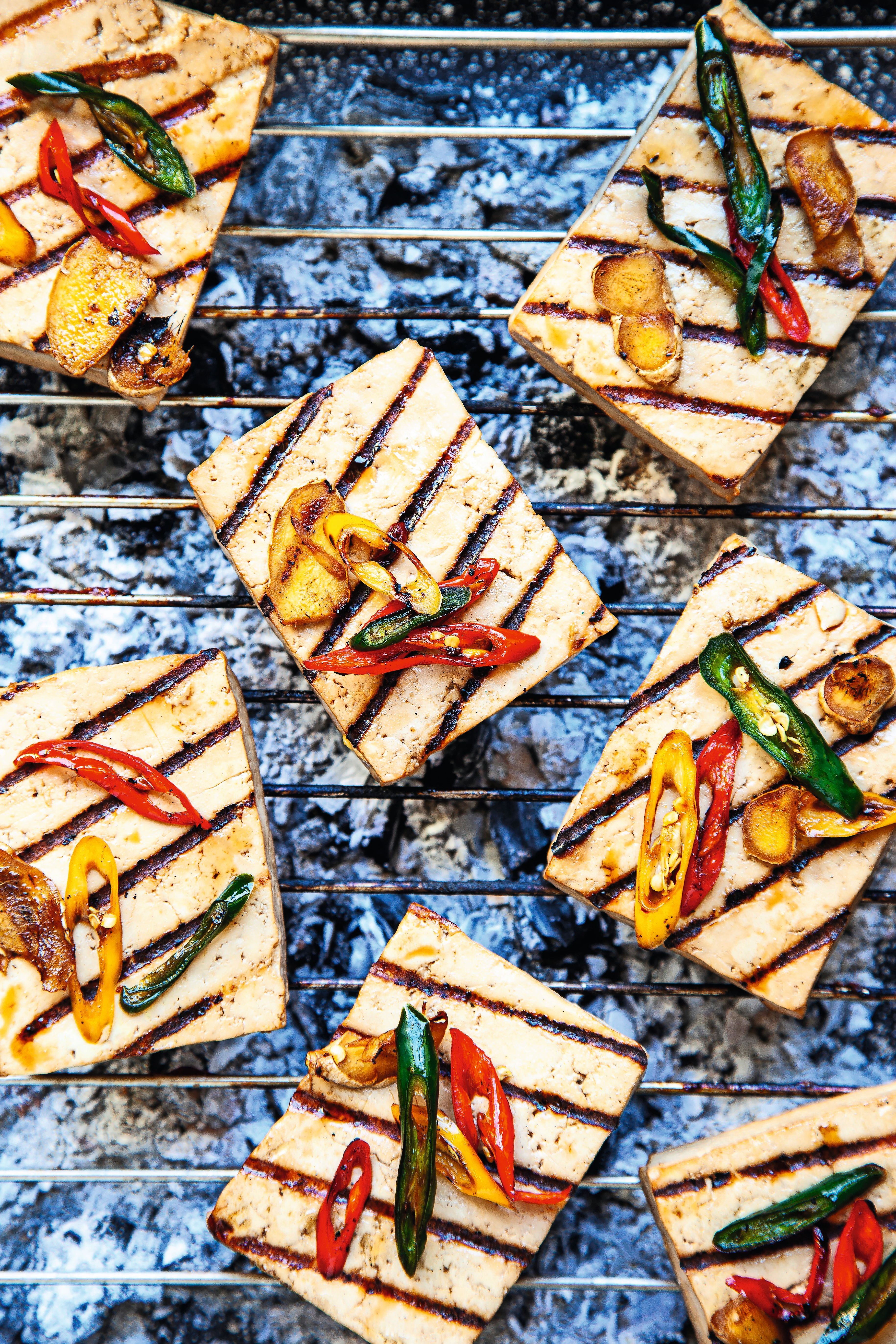 Gegrillter Tofu mit bunten Chilis und Ingwerscheiben auf einem Rost über Holzkohle.