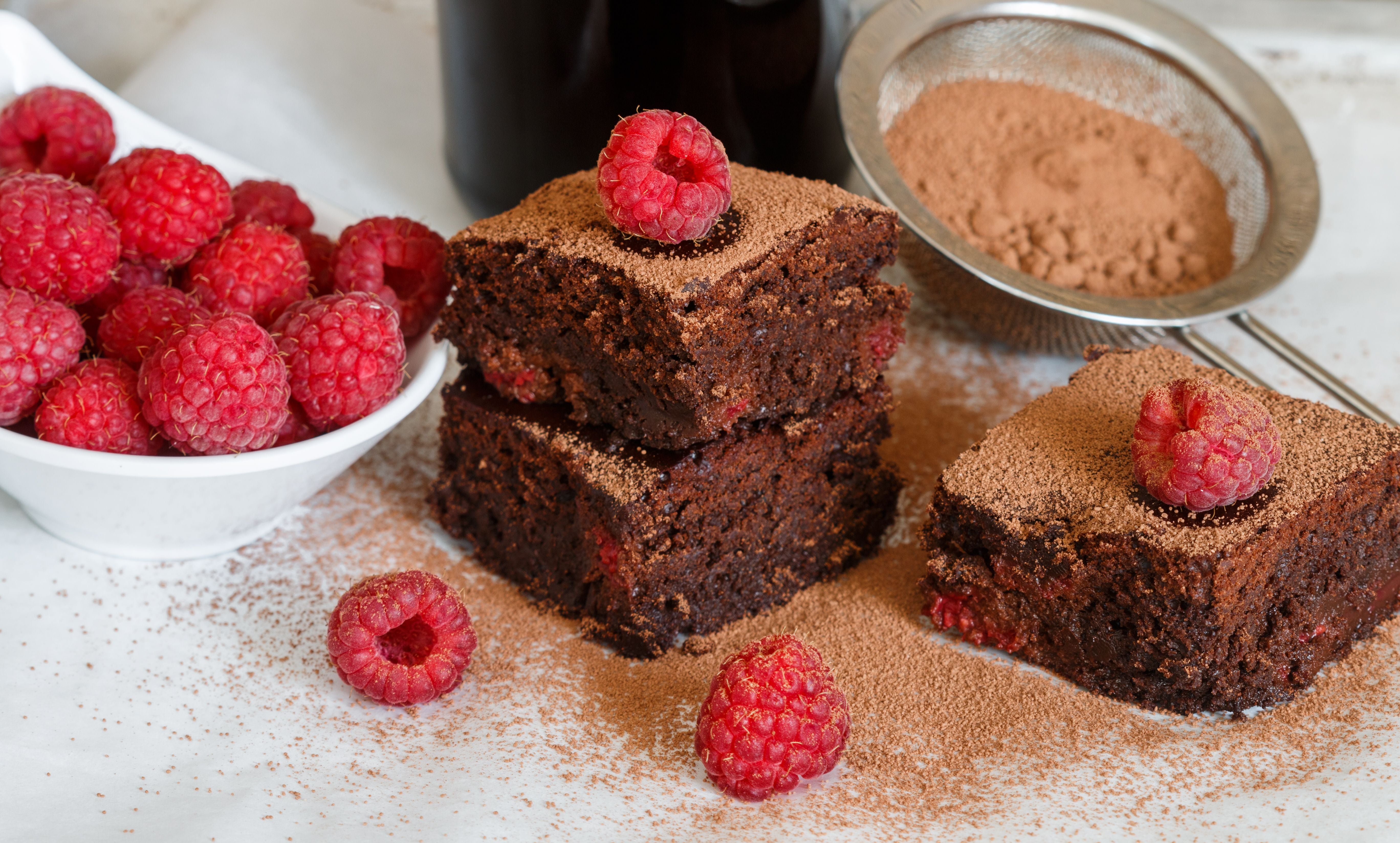 Drei Schokoladen-Brownies mit Himbeeren und Kakaopulver neben frischen Himbeeren.