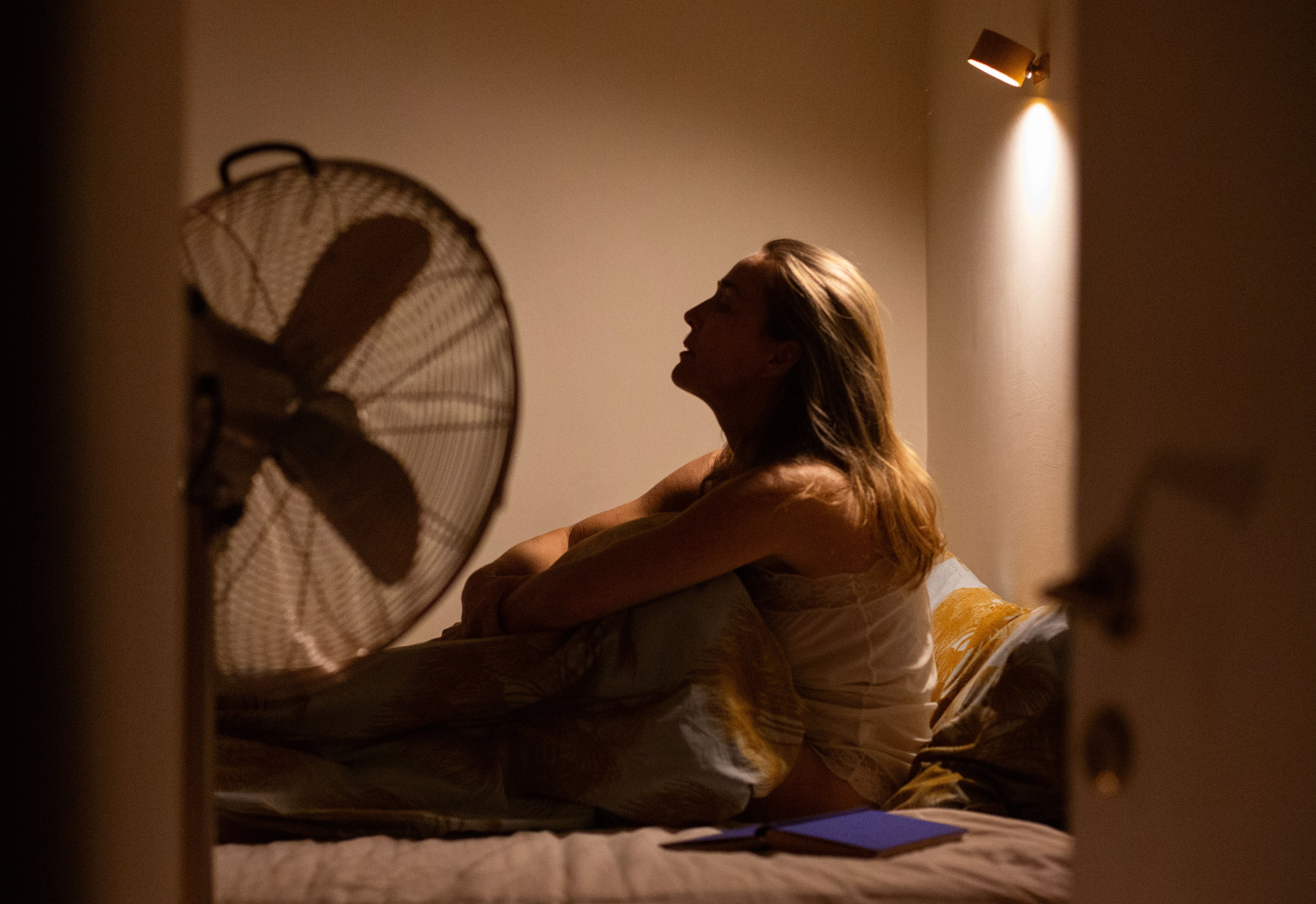 Frau sitzt nachts nachdenklich im Bett, neben ihr ein Ventilator und eine Lampe an der Wand.