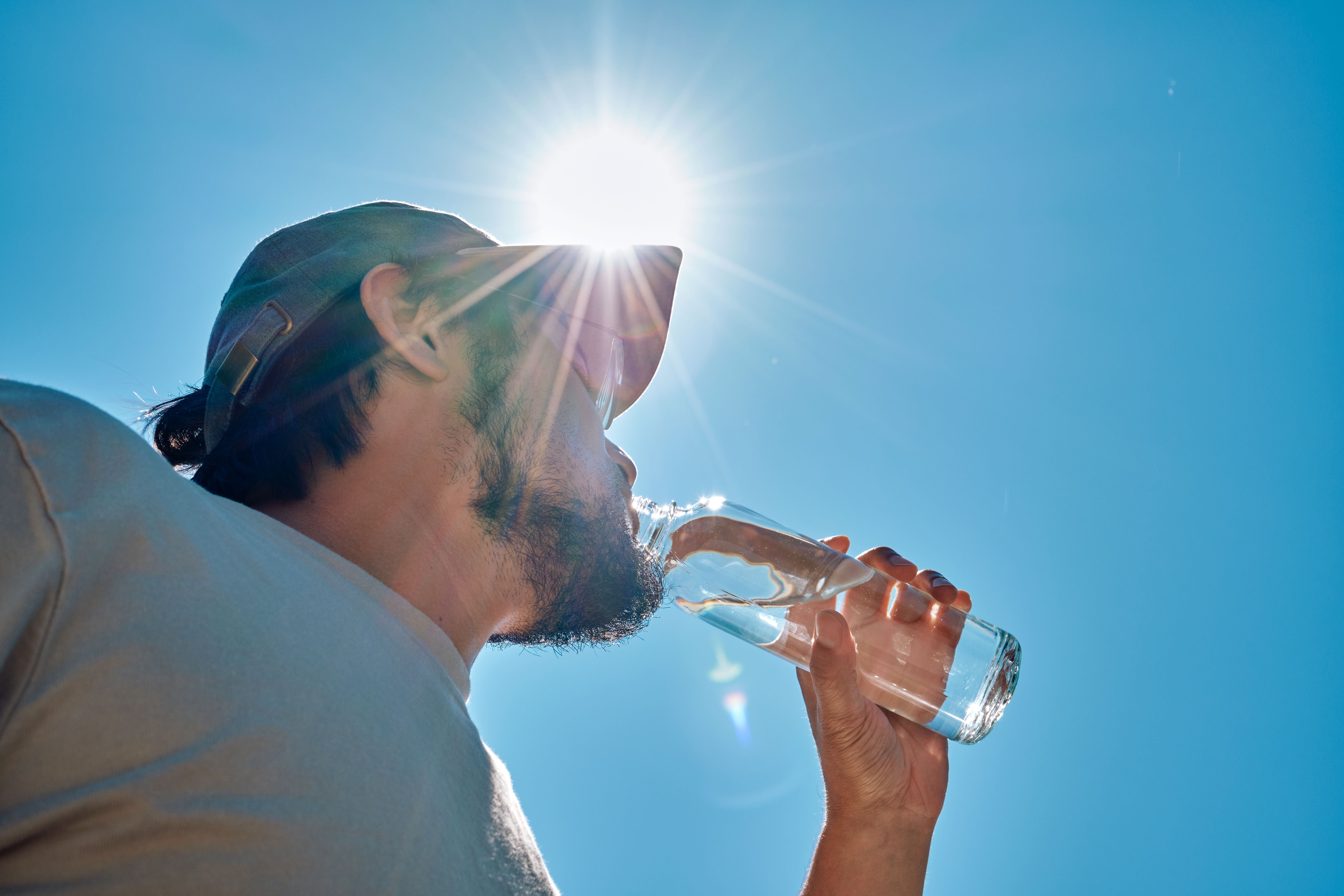 Mann mit Kappe trinkt bei Sonnenschein aus einer Wasserflasche vor blauem Himmel.