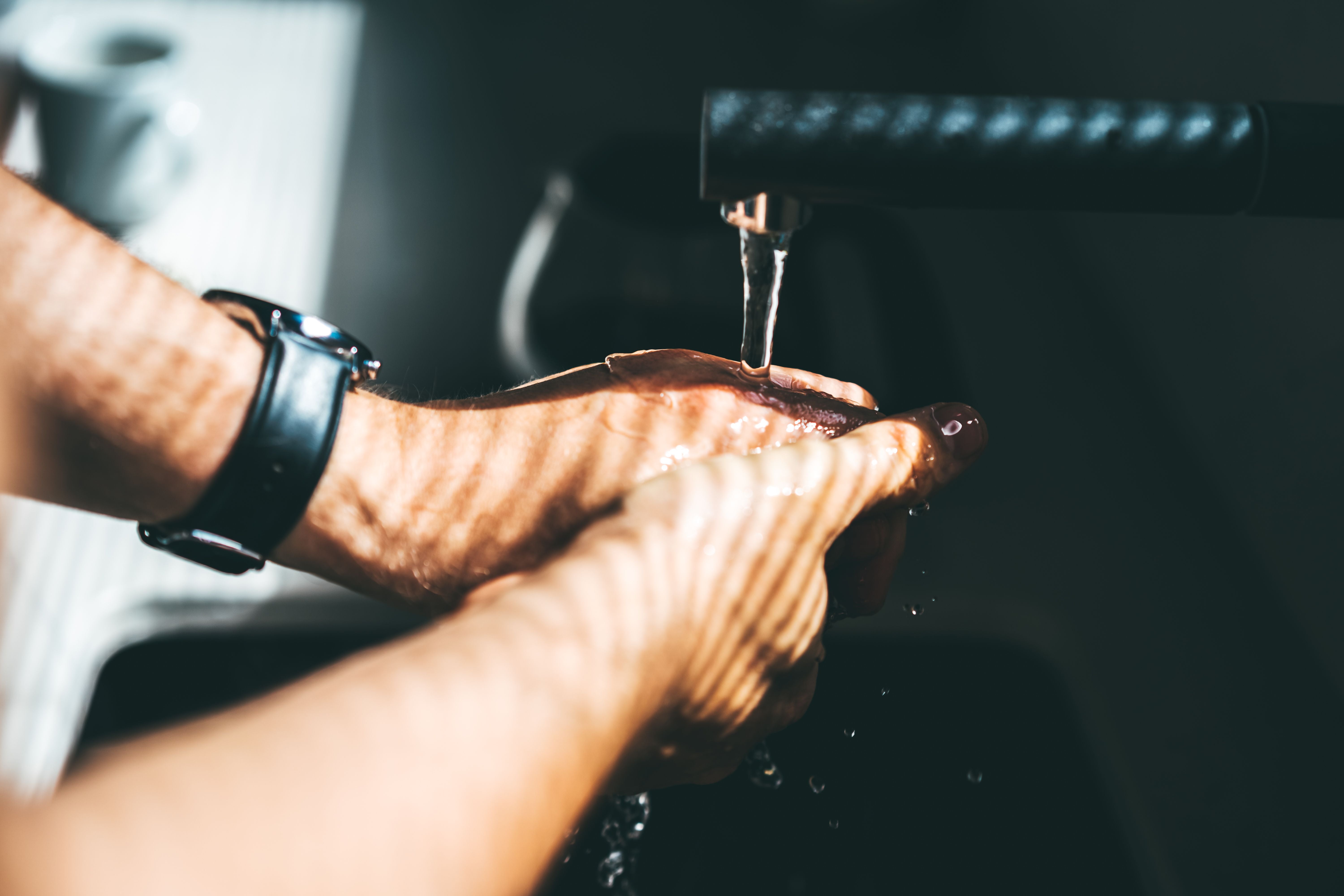 Mann mit Uhr wäscht sich bei Sonnenlicht die Hände unter fließendem Wasser am Waschbecken.