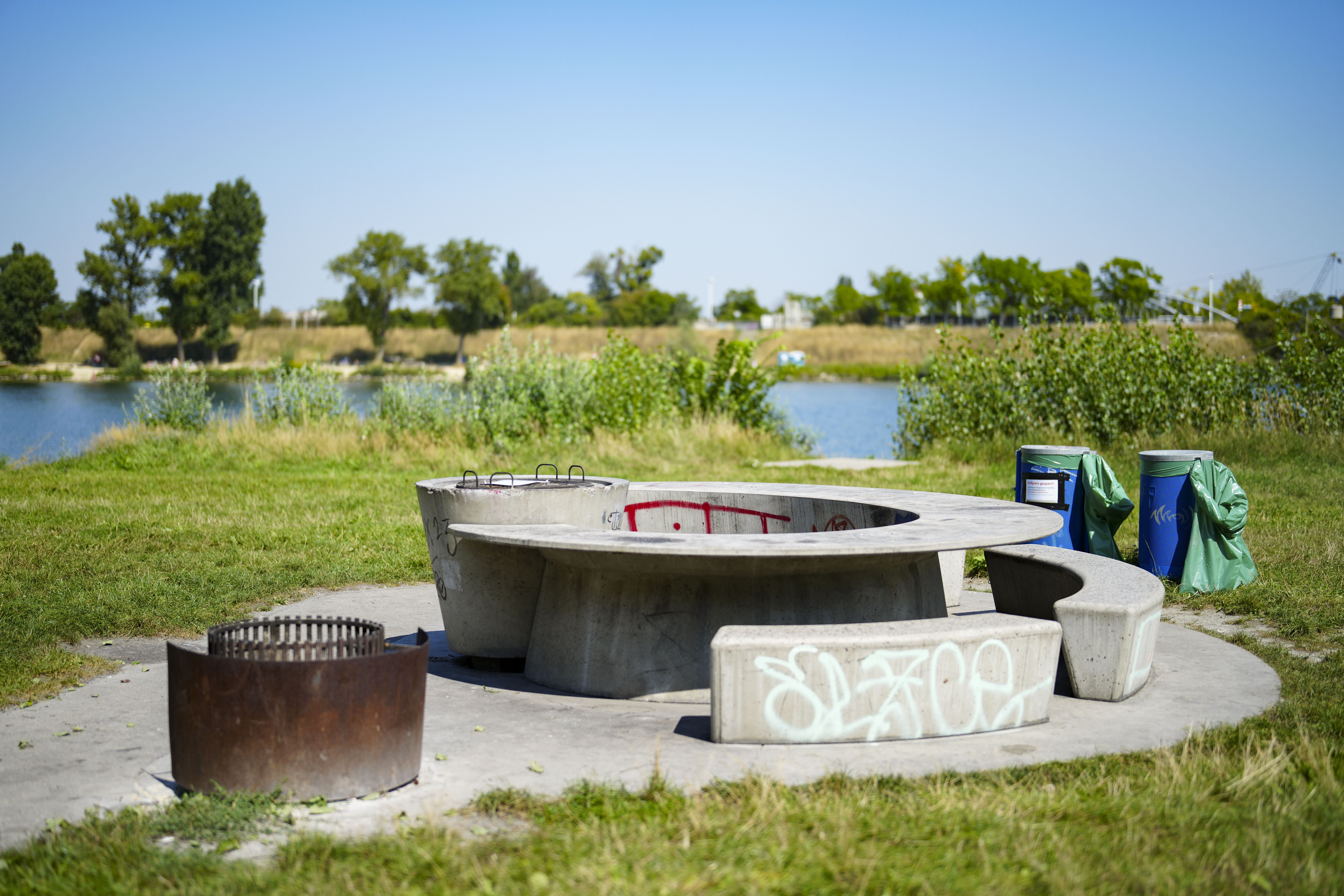 Gesperrter Grillplatz mit Sitzbank und Abfalleimern auf der Donauinsel in Wien bei sonnigem Wetter.