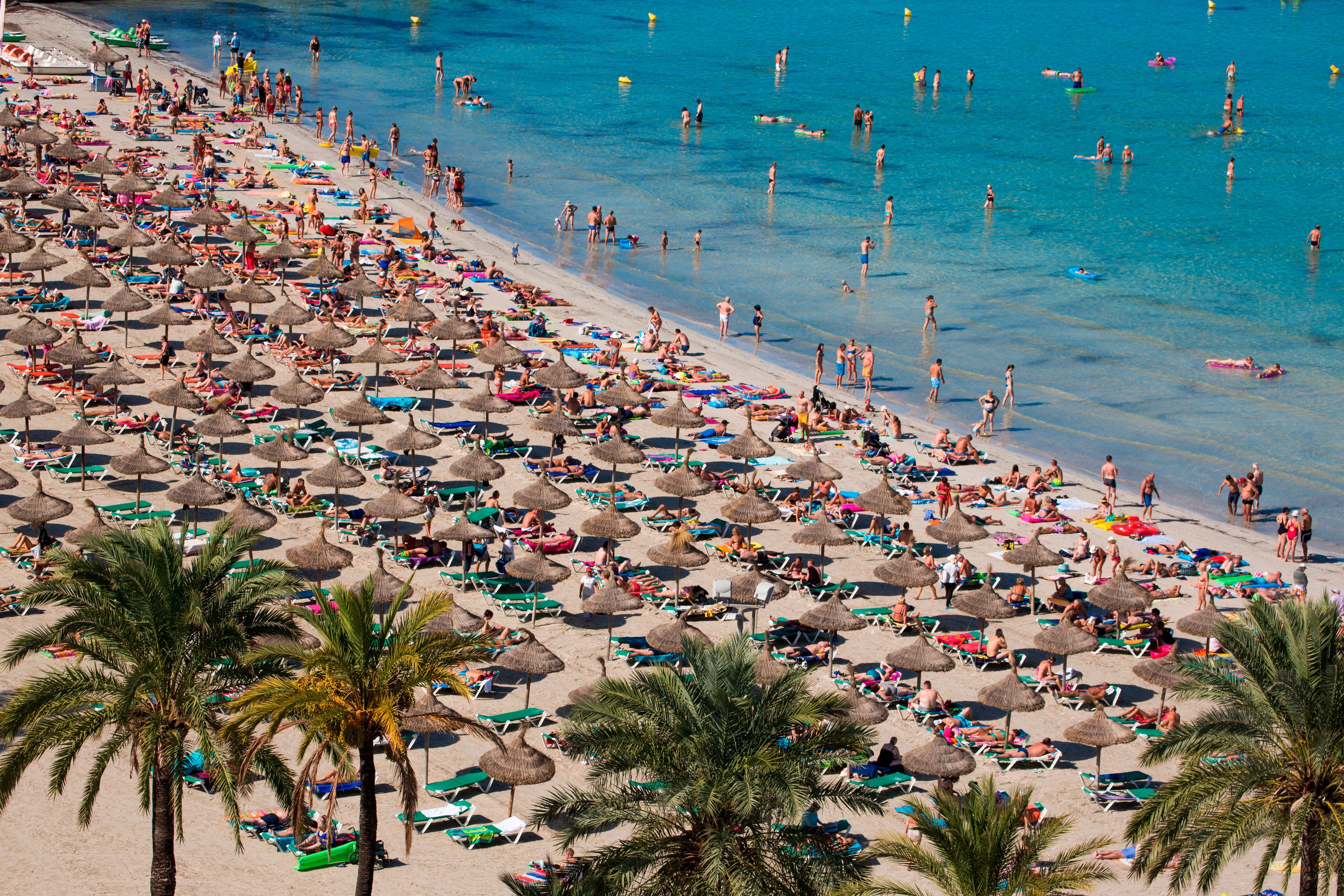 Viele Menschen entspannen unter Sonnenschirmen am vollen Sandstrand mit Palmen und klarem Meer.