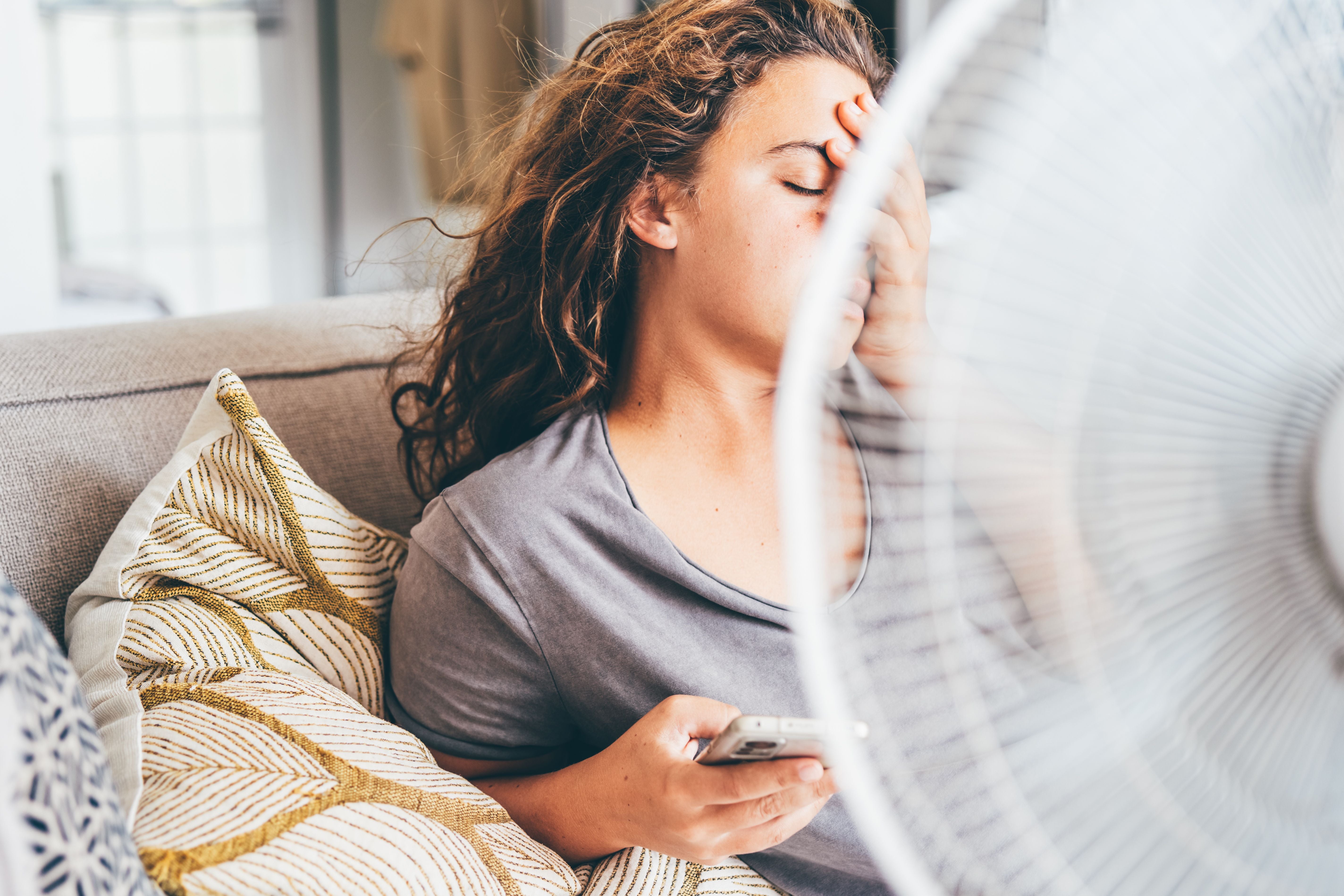Frau sitzt erschöpft vor Ventilator auf dem Sofa und hält ein Handy in der Hand.