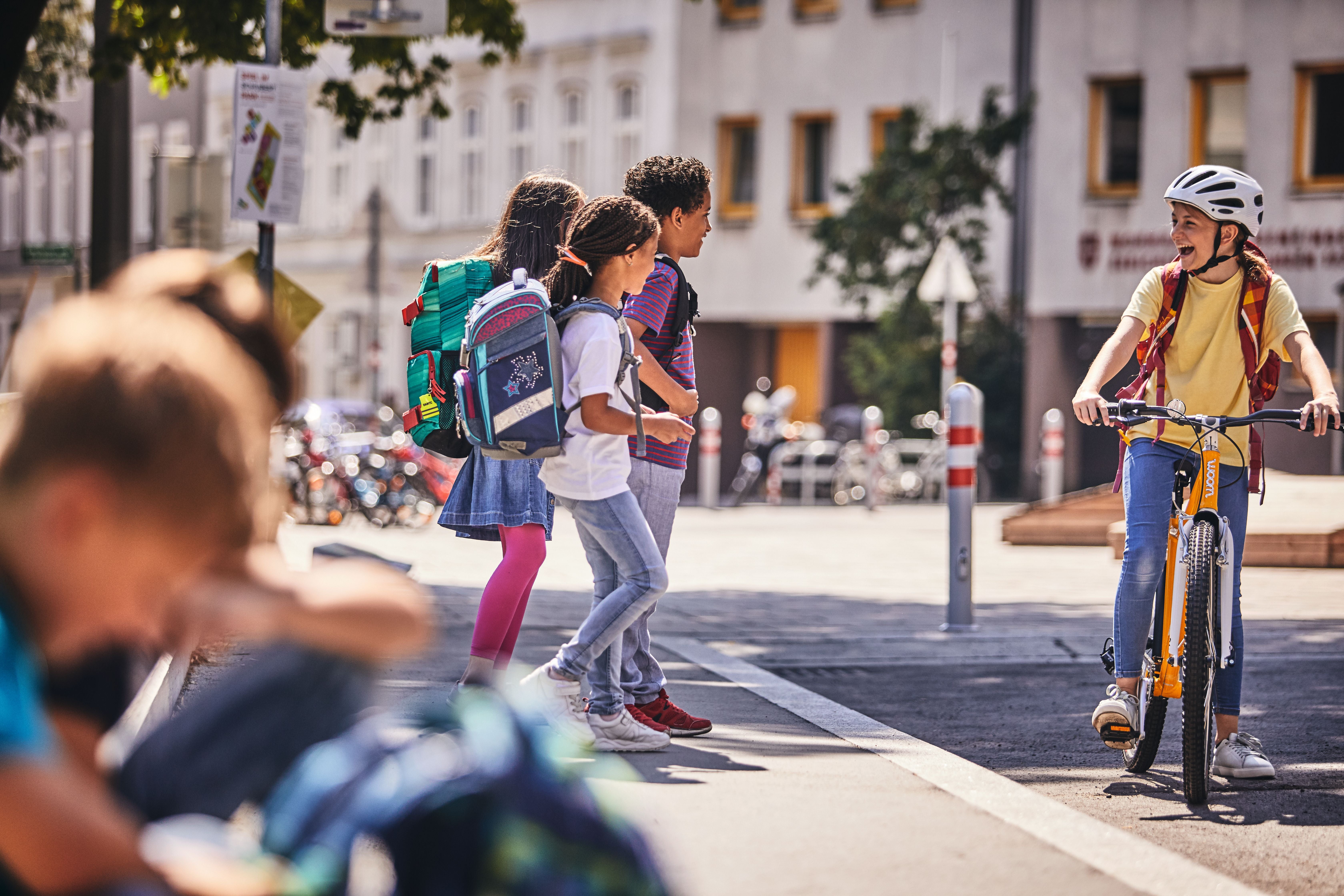 Kinder mit Rucksäcken gehen über die Straße, ein Mädchen mit Fahrrad fährt lachend vorbei.