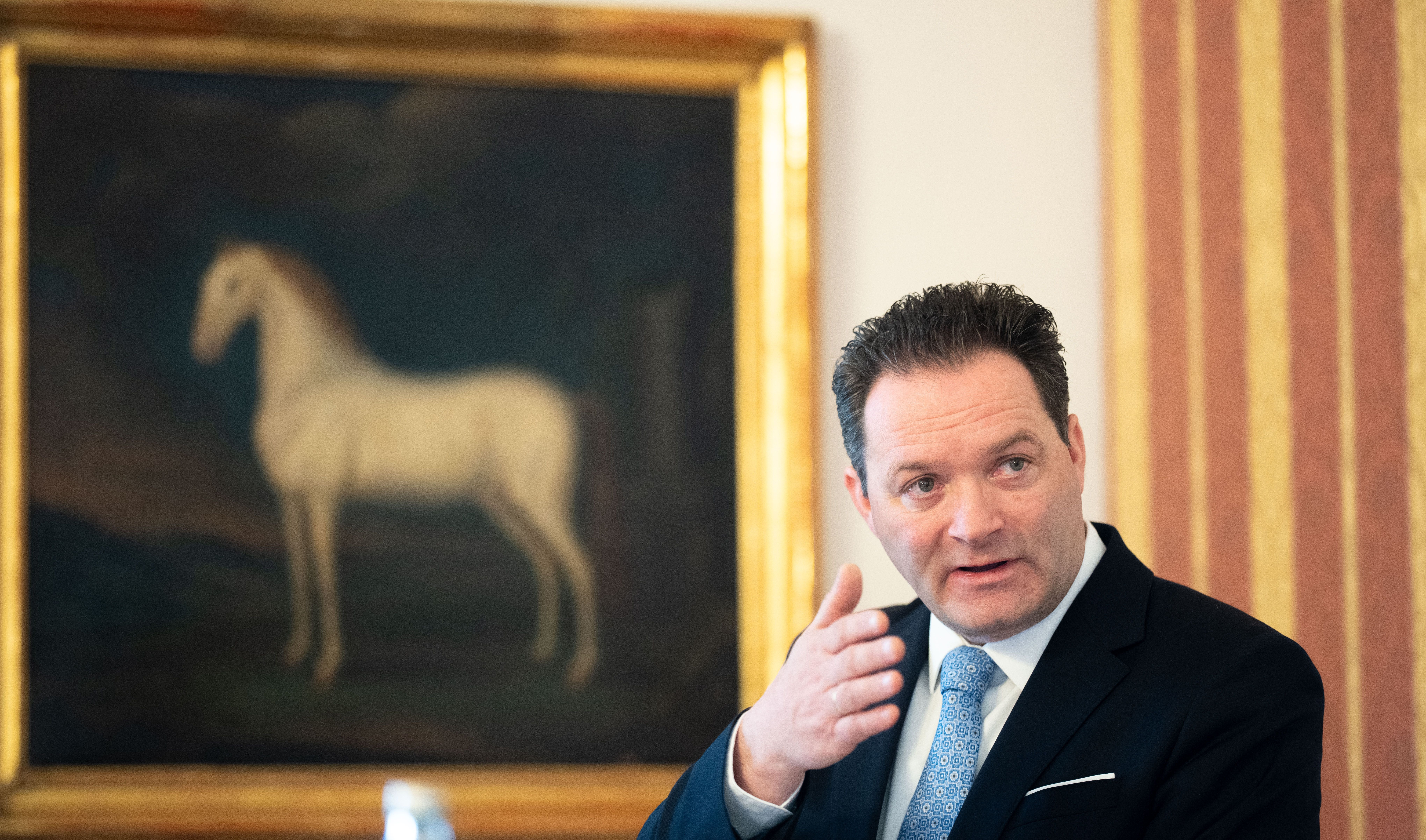 Ein Mann im Anzug spricht, im Hintergrund ein Gemälde eines weißen Pferdes.