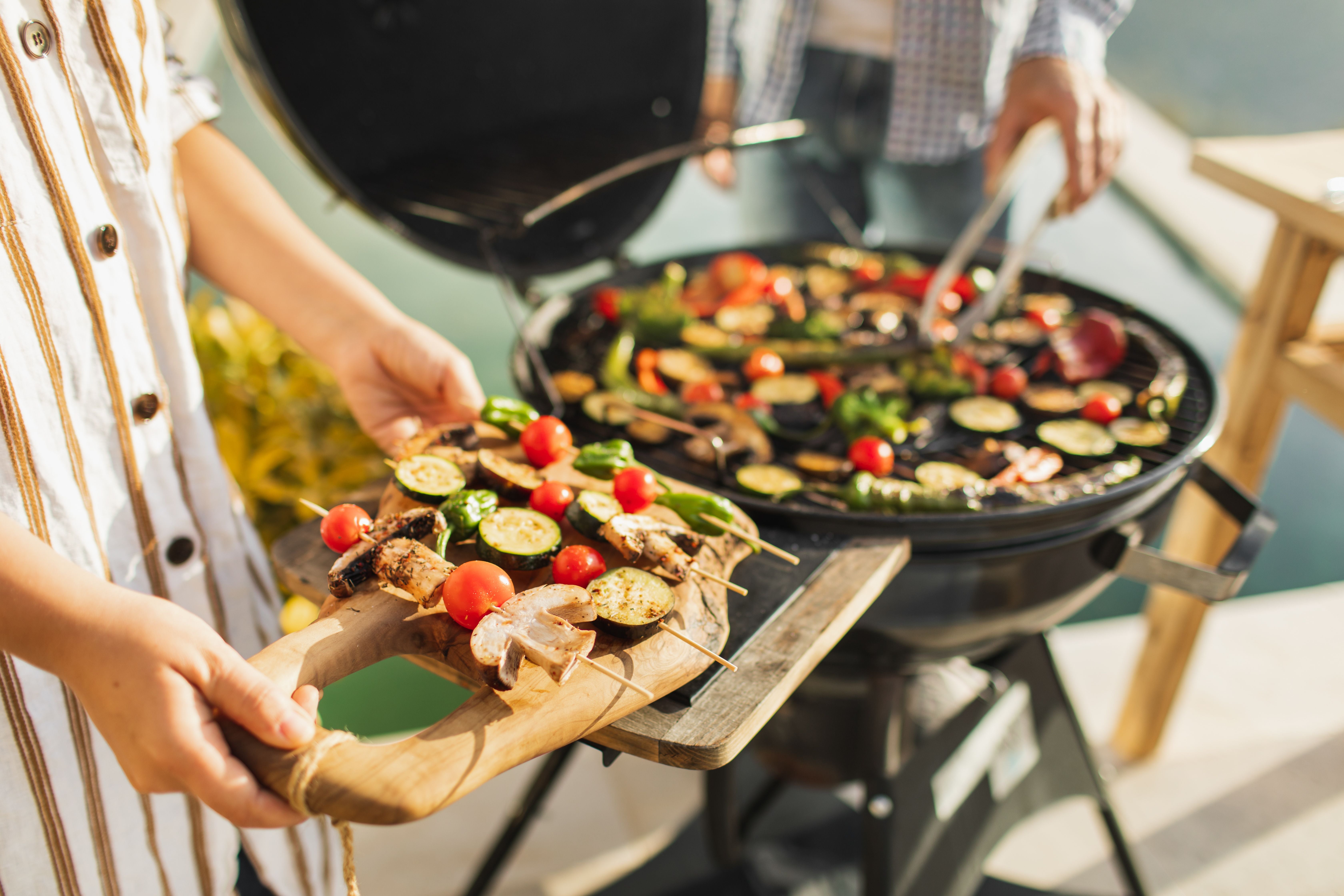 Gemüsespieße werden auf einem Grill im Freien zubereitet, zwei Personen sind beteiligt.