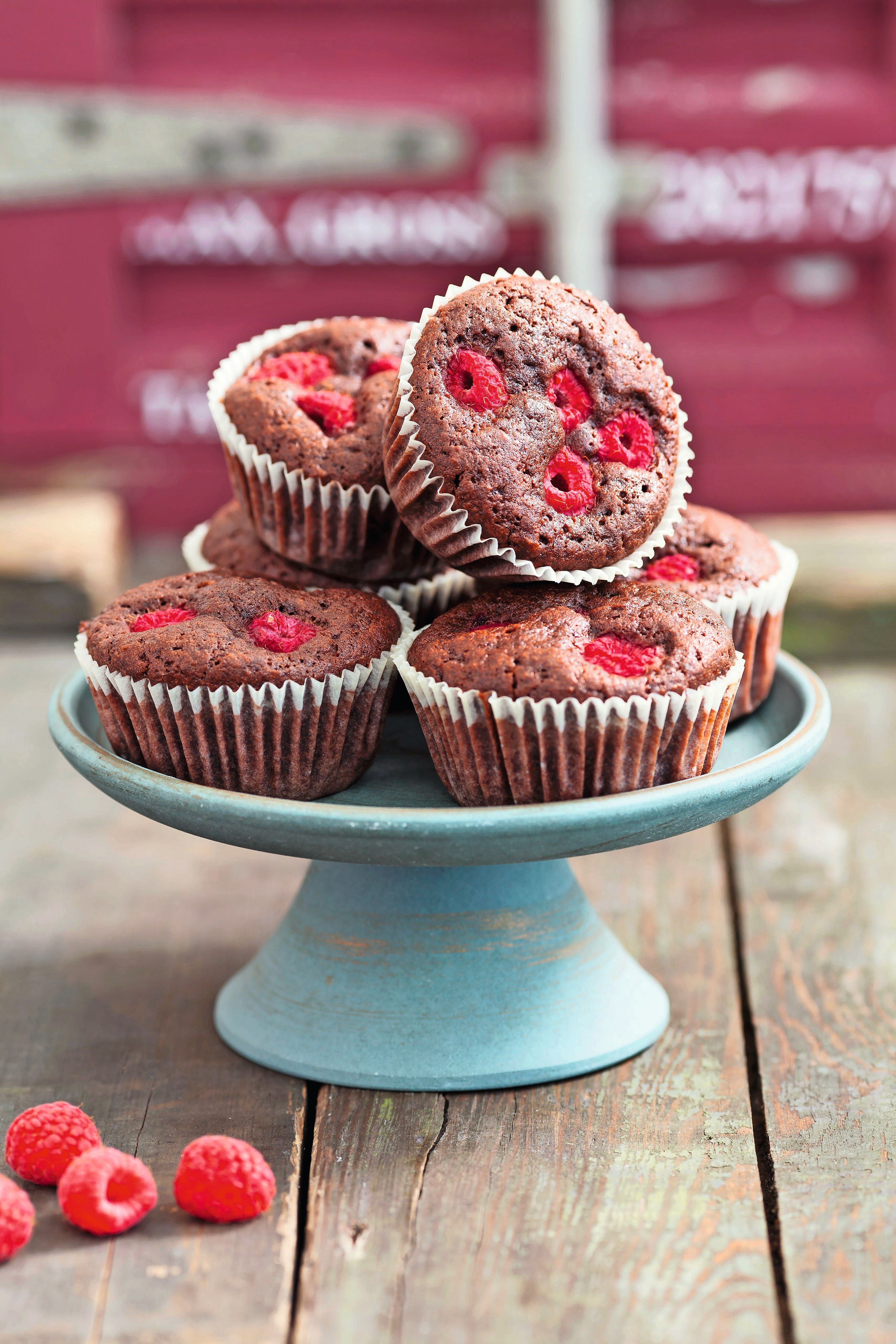 Sechs Brownie-Muffins mit Himbeeren auf einer grünen Tortenplatte, im Vordergrund frische Himbeeren.