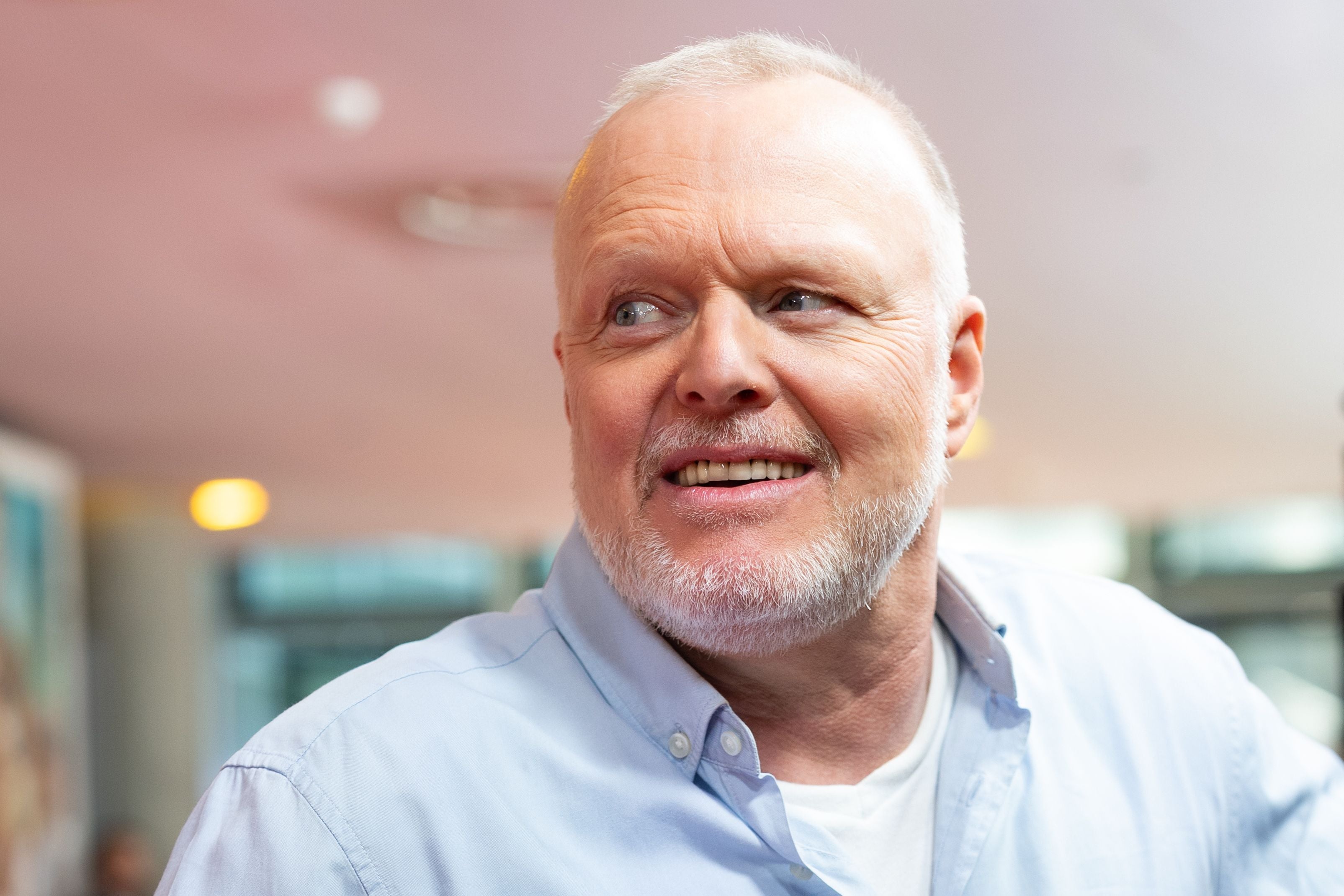 Ein älterer Mann mit grauem Bart lächelt und blickt zur Seite, trägt ein hellblaues Hemd.