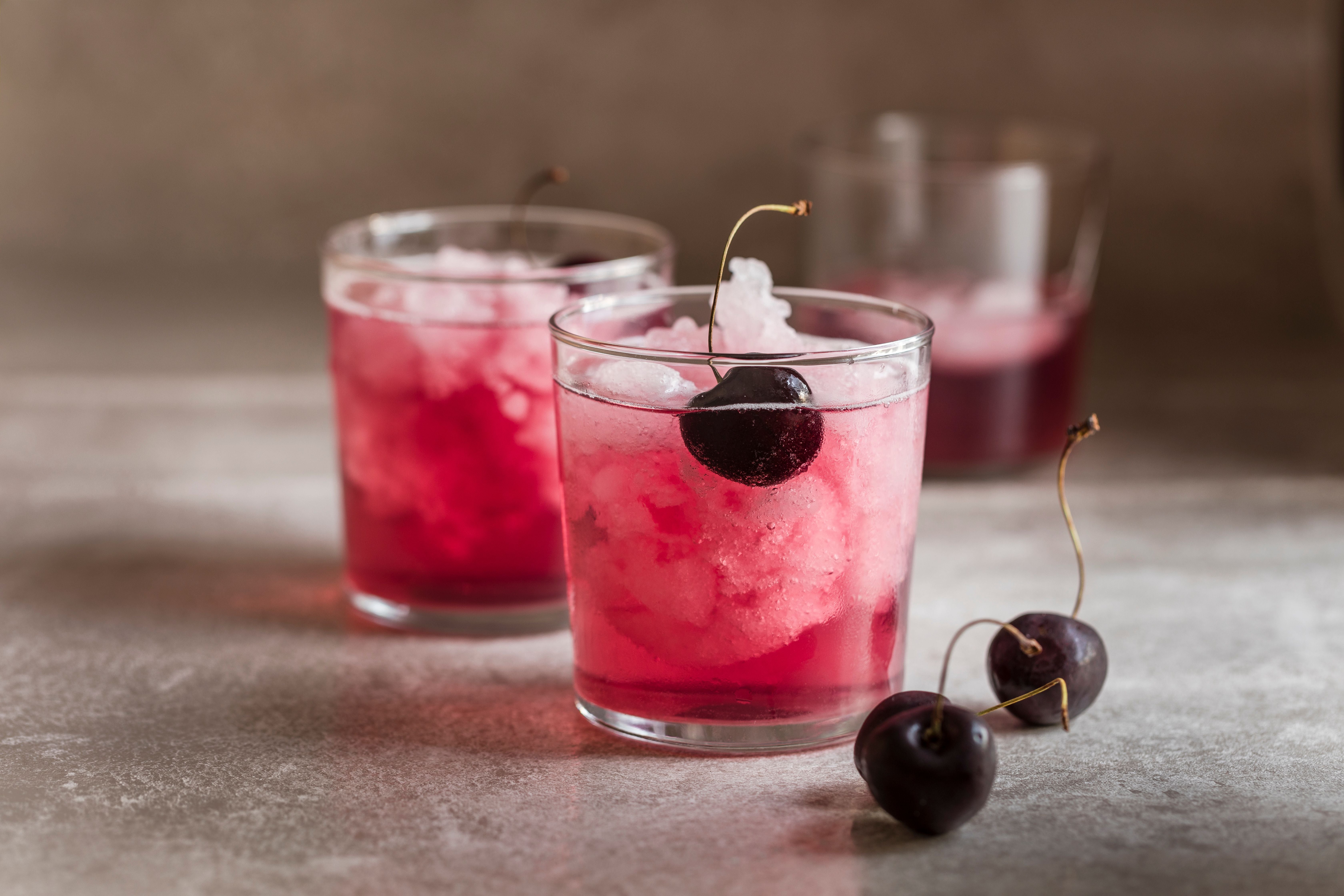 Rotes Getränk mit Eis und Schwarzkirsche im Glas, zwei Kirschen liegen daneben, weitere Gläser im Hintergrund.