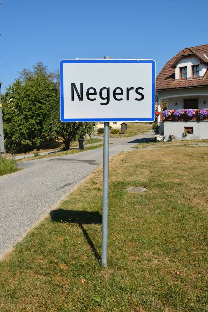 Ortsschild mit dem Namen "Negers" vor einem Haus und einer Nebenstraße im Grünen.