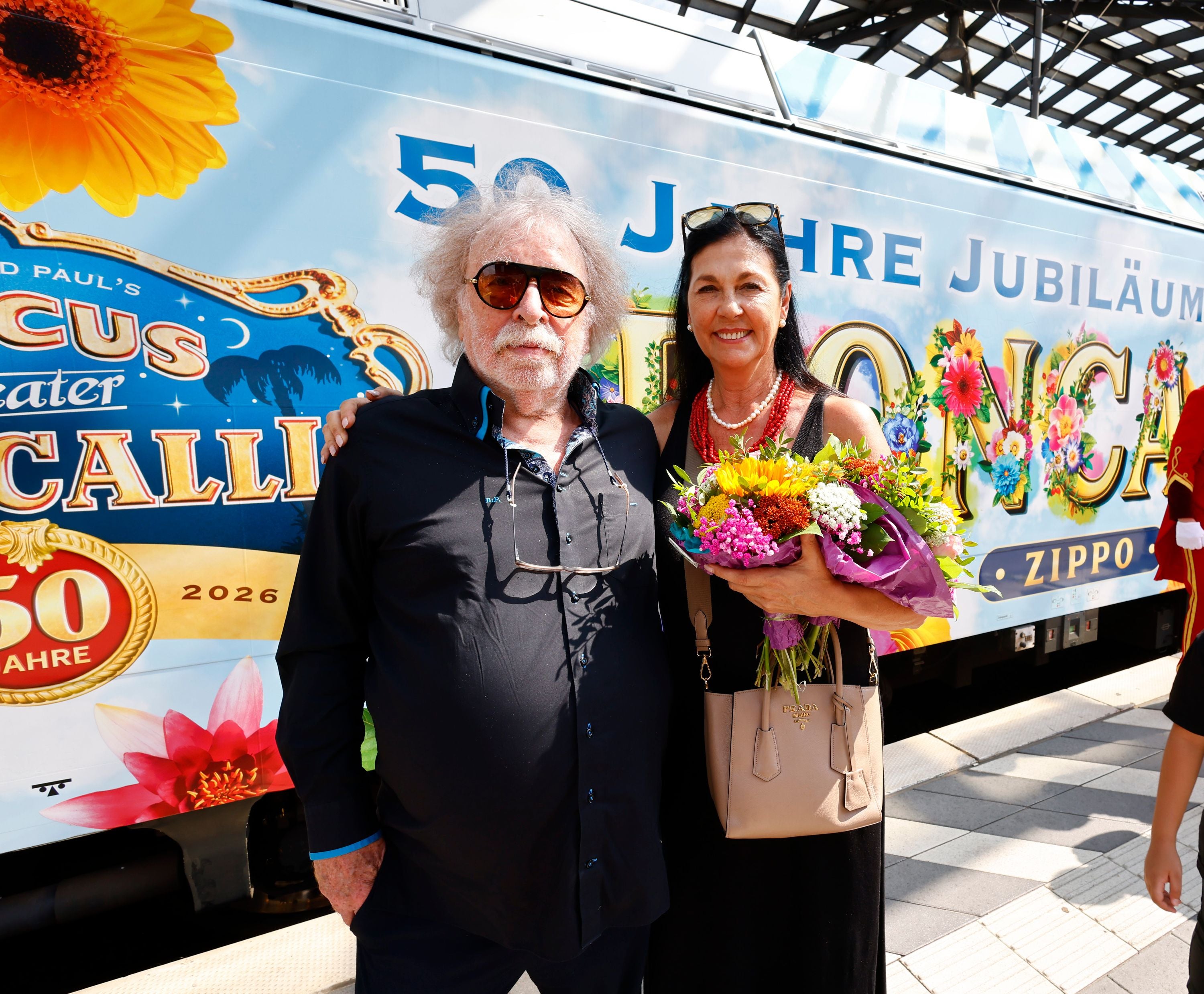 Ein Mann und eine Frau stehen lächelnd vor einem bunt bemalten Roncalli-Zug, die Frau hält Blumen.