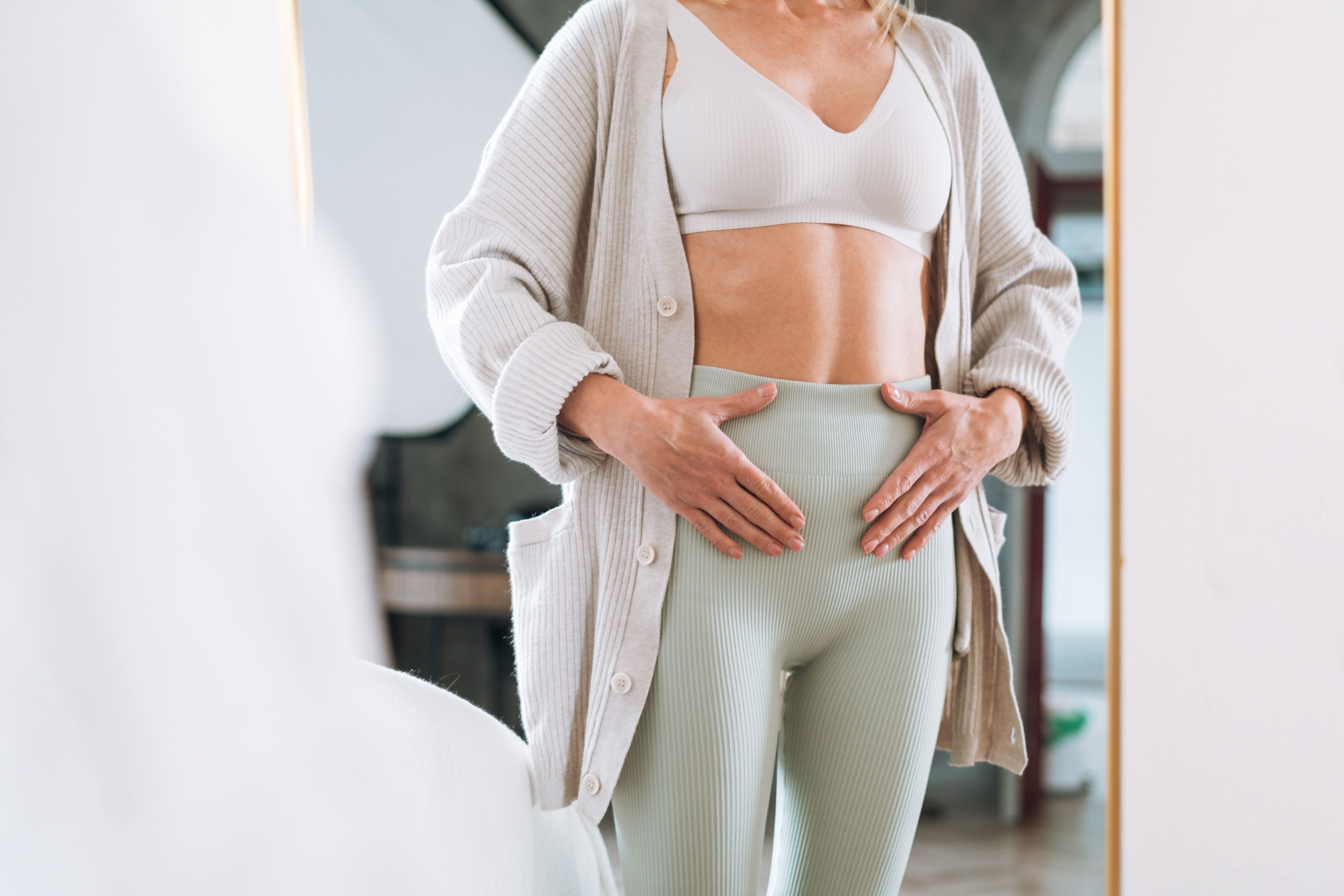 Frau steht vor dem Spiegel und hält sanft ihren Unterbauch, Symbol für Frauengesundheit und Wohlbefinden.