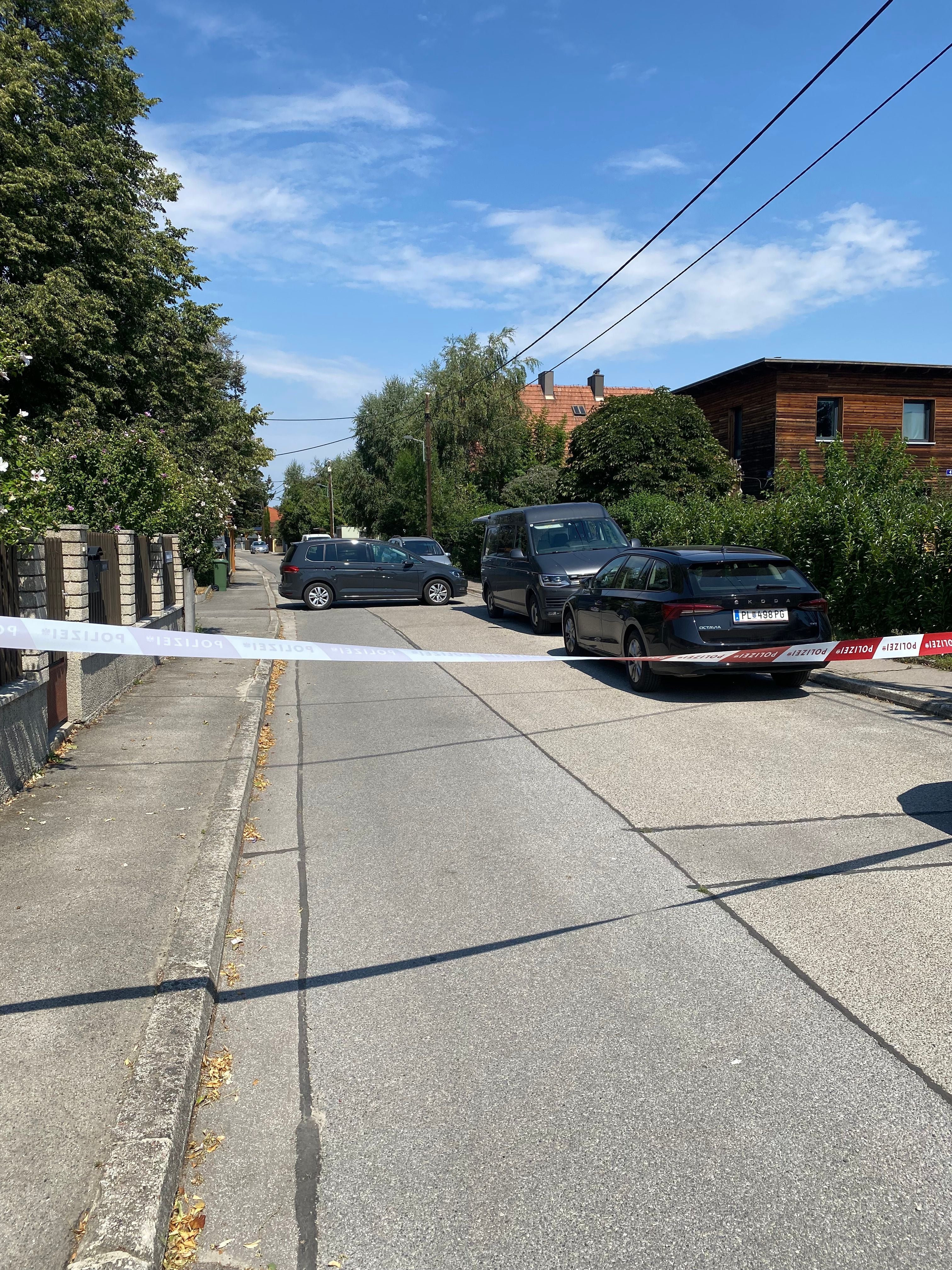 Polizeiabsperrung blockiert Straße in Wohngebiet, mehrere geparkte Autos, sonniges Wetter.