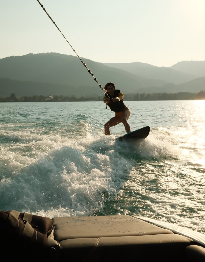 Person surft hinter einem Boot bei Sonnenuntergang auf einem See, Berge im Hintergrund.