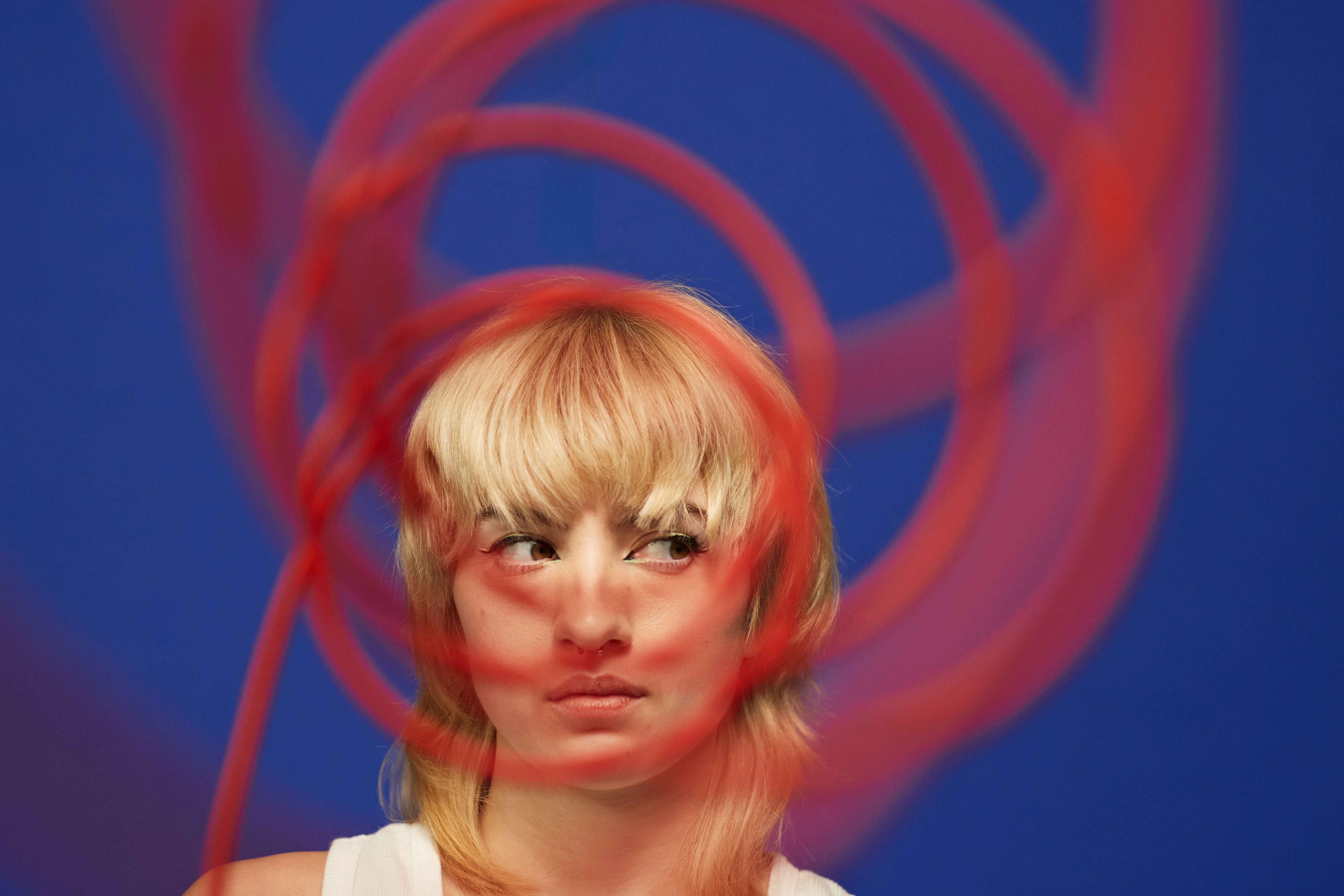 Junge nicht-binäre Person mit blondem Haar blickt hinter rotem Spiralkabel vor blauem Hintergrund seitlich.