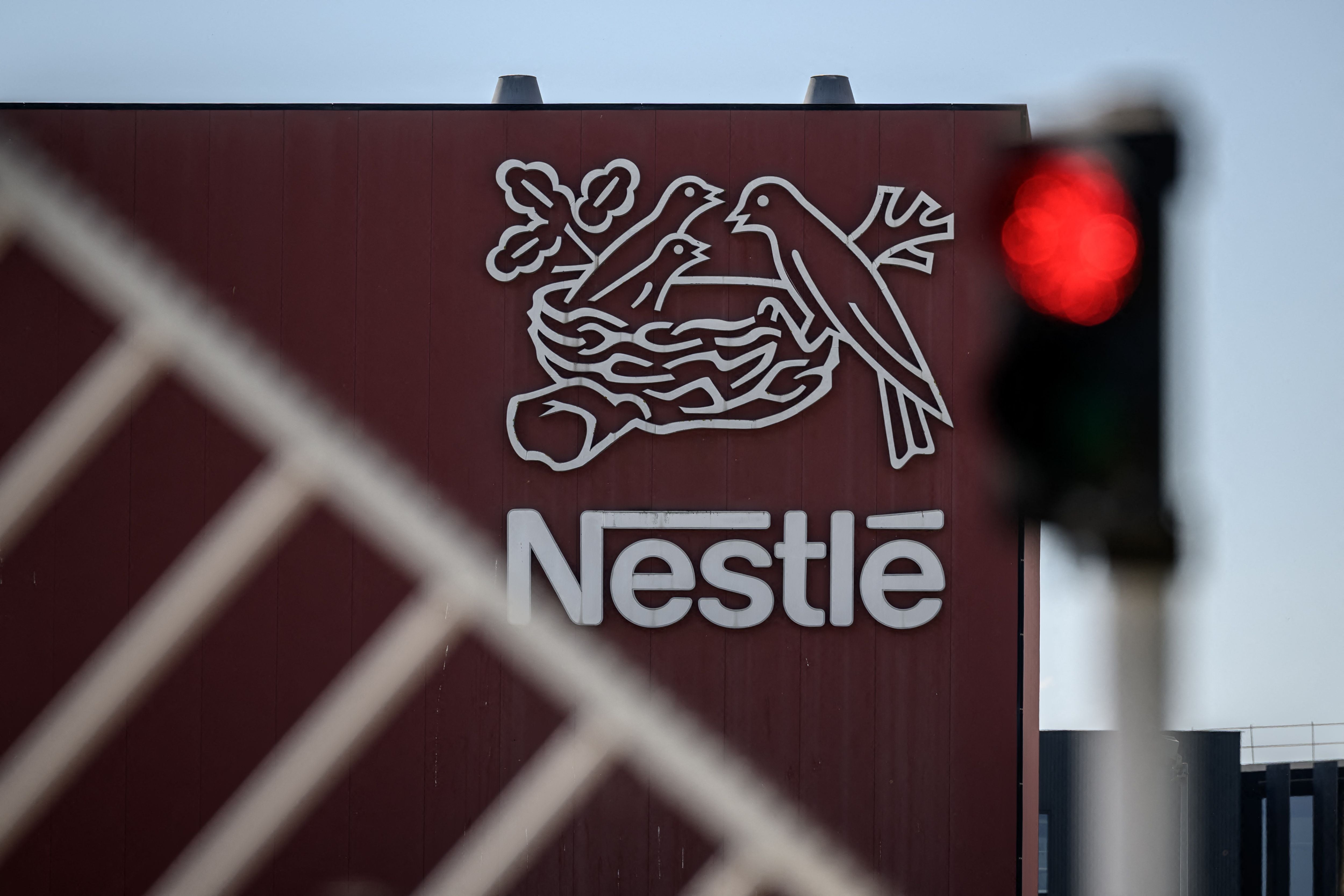 Nestlé-Logo an einer Fabrikfassade, vorne unscharf ein Zaun und eine rote Ampel.