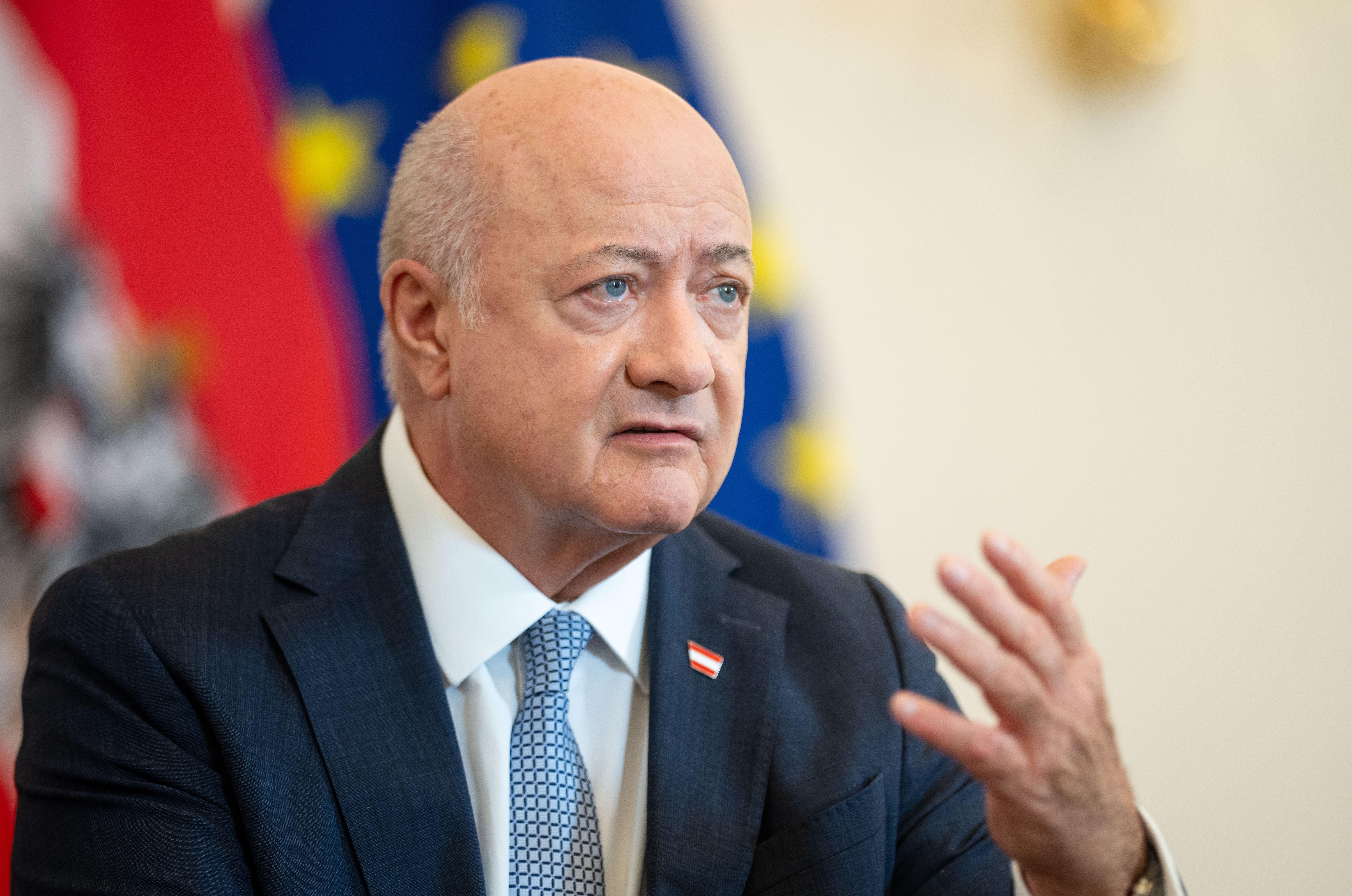 Ein Mann im Anzug spricht und gestikuliert vor verschwommenen Fahnen im Hintergrund.
