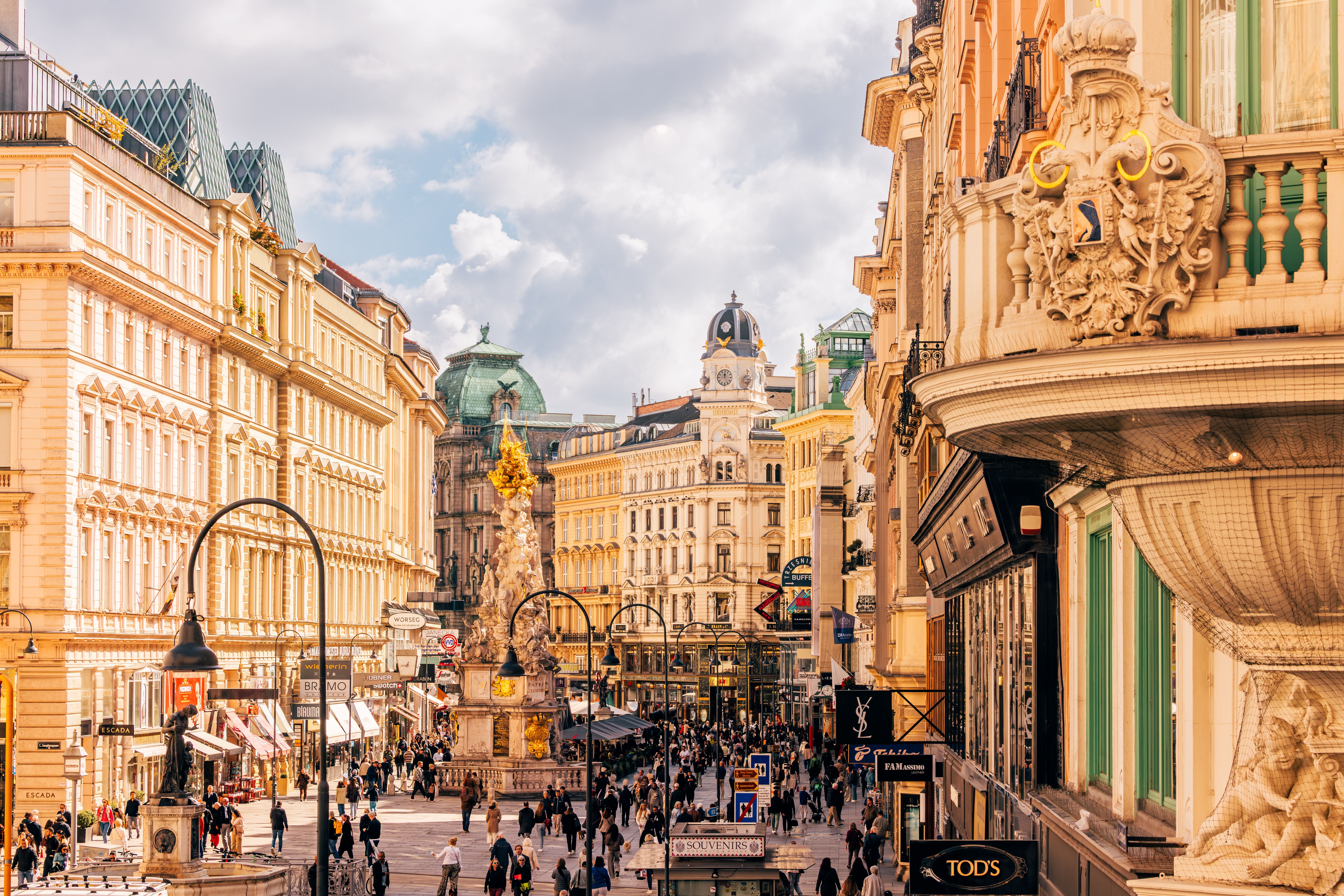 Belebte Einkaufsstraße in Wien mit historischen Gebäuden und goldener Pestsäule bei Tageslicht.