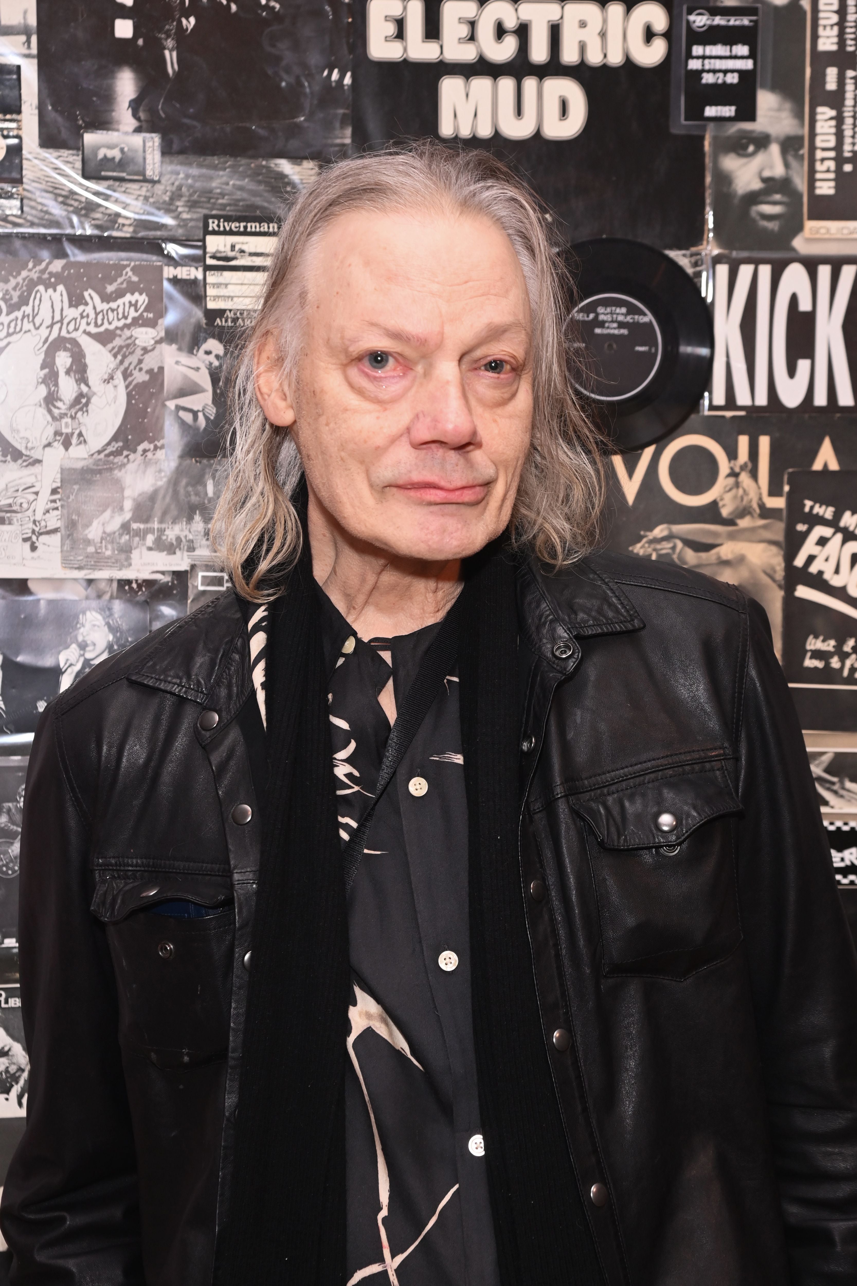 Ein älterer Mann mit langen Haaren steht vor einer Wand mit Rockmusik-Postern und trägt Lederjacke.
