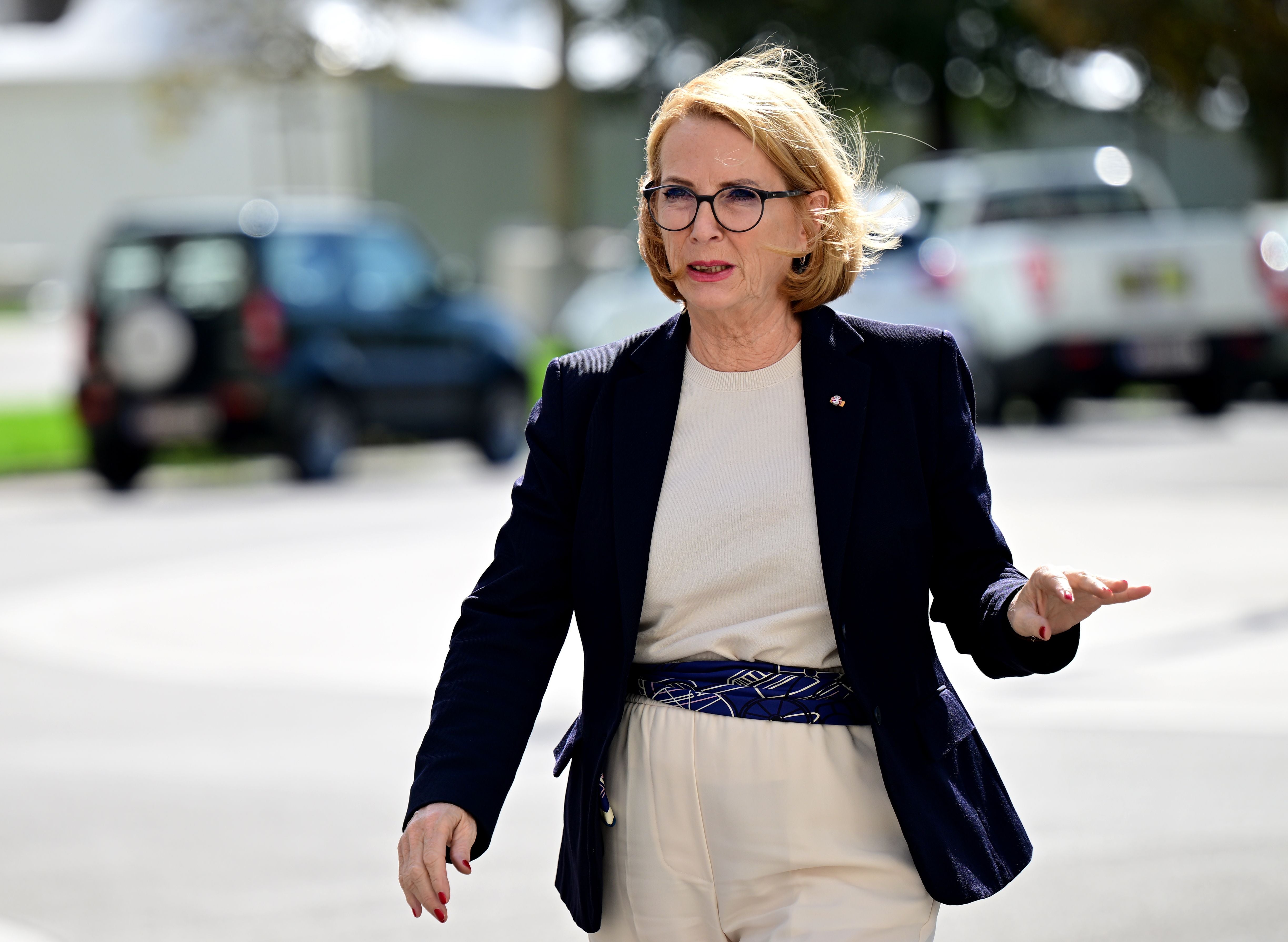 Eine Frau mit Brille und blondem Haar geht im Freien, sie trägt Blazer und helle Hose.