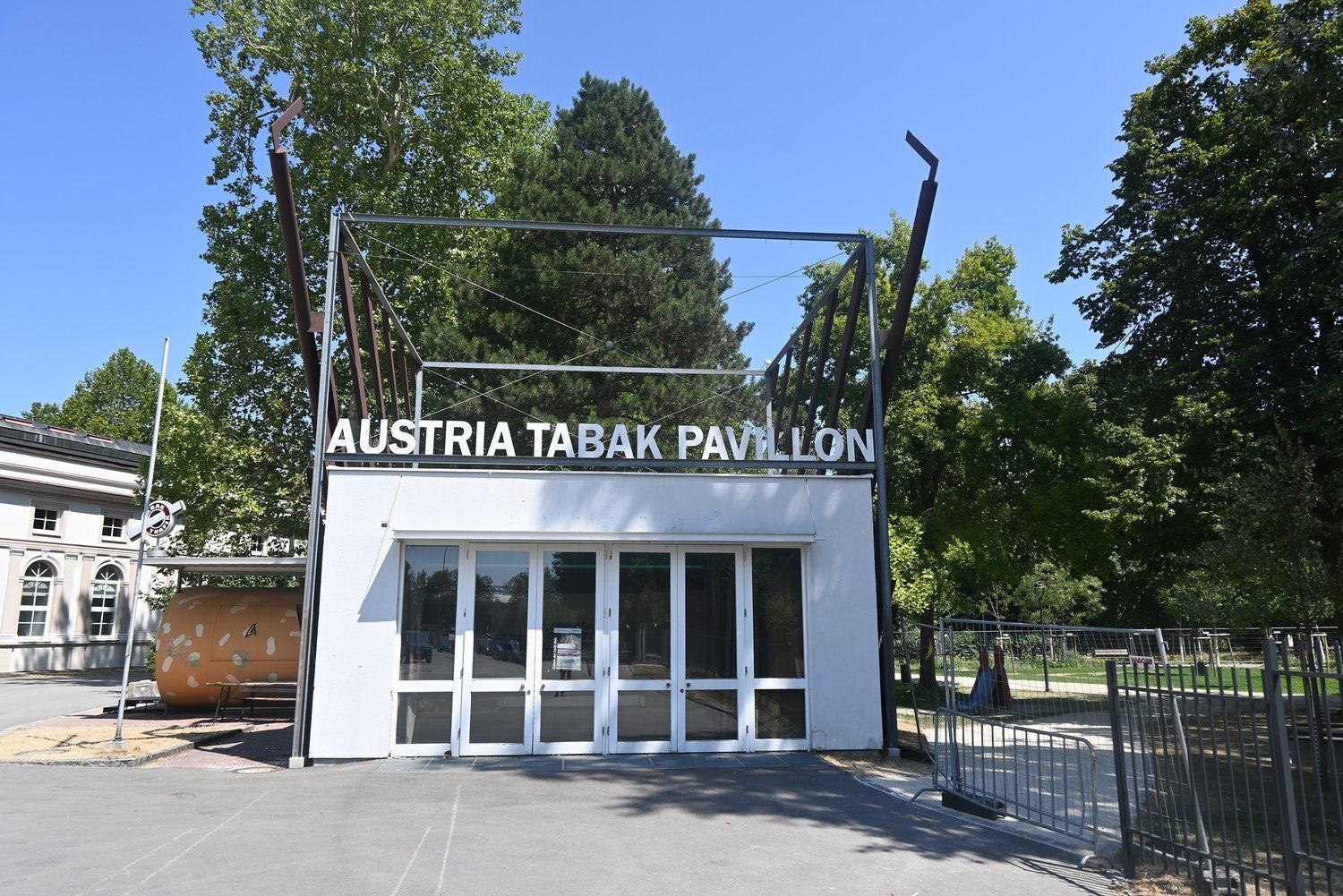 Moderner weißer Pavillon mit Aufschrift "AUSTRIA TABAK PAVILLON" vor Bäumen und blauem Himmel.