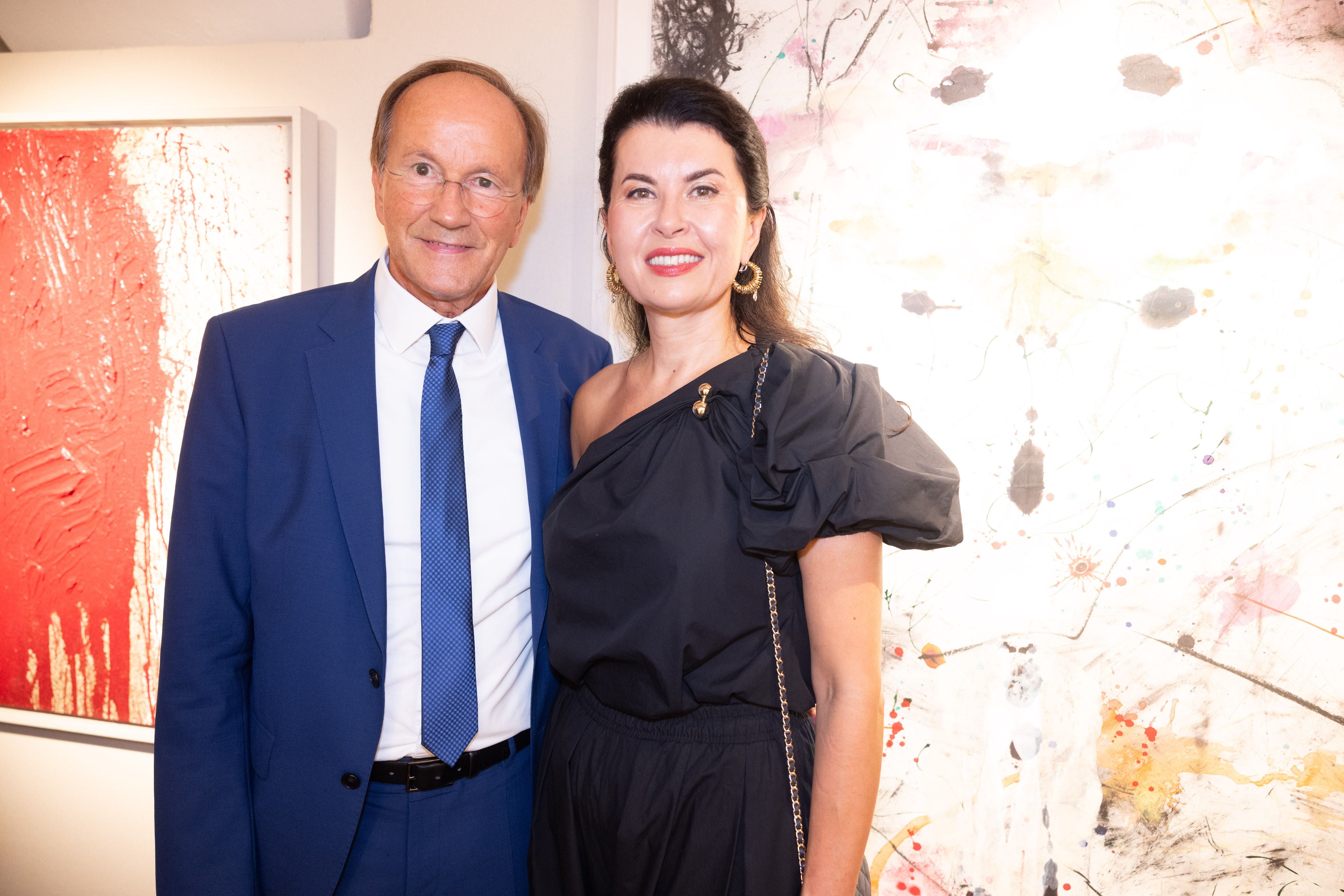 Ein Mann im blauen Anzug und eine Frau im schwarzen Kleid lächeln vor abstrakter Kunst.