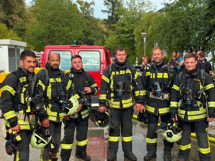 Sechs Feuerwehrmänner in Schutzanzügen stehen vor einem roten Einsatzfahrzeug im Freien.