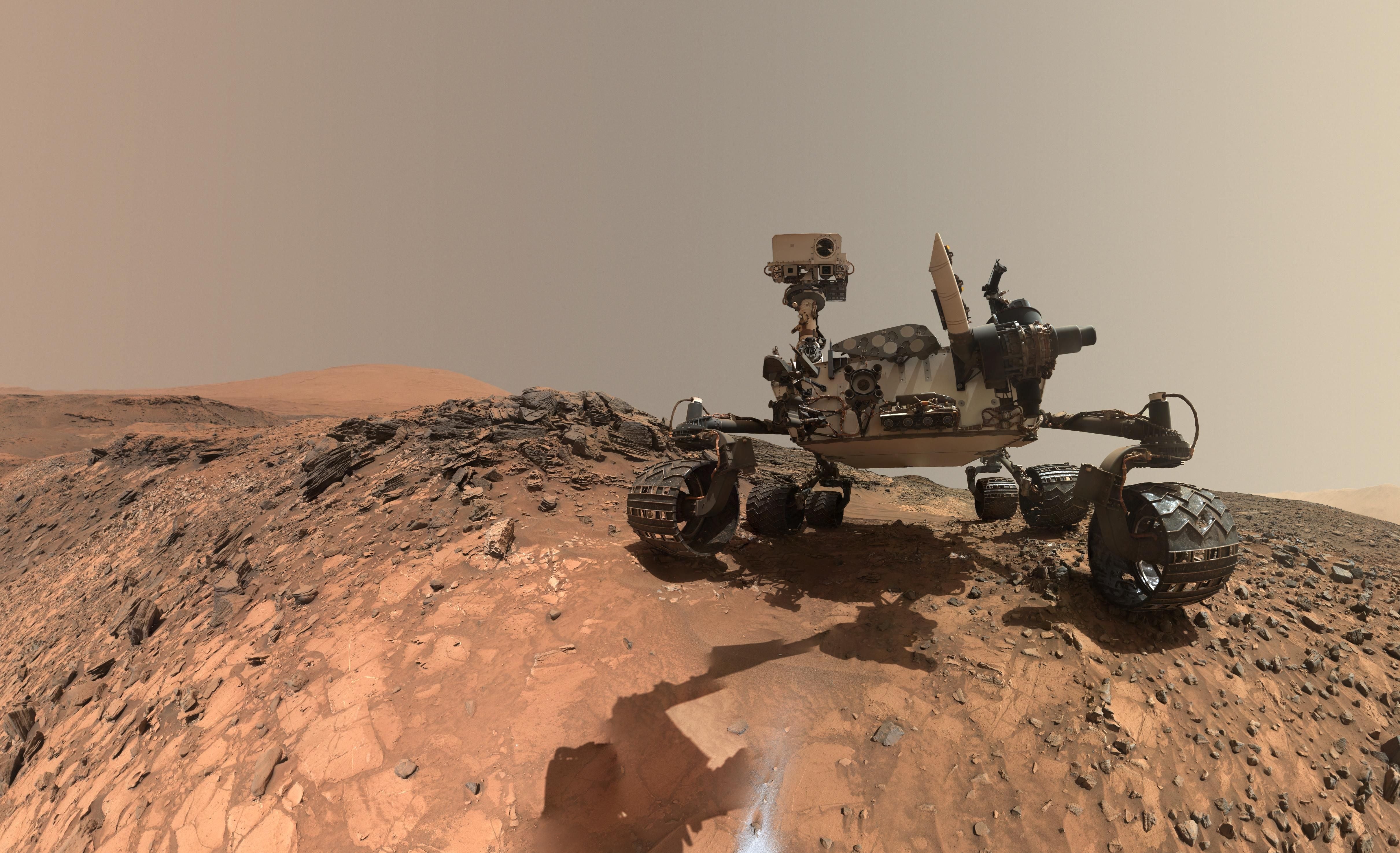 Marsrover Curiosity macht ein Selfie auf felsigem Gelände am Mount Sharp unter rötlichem Himmel.