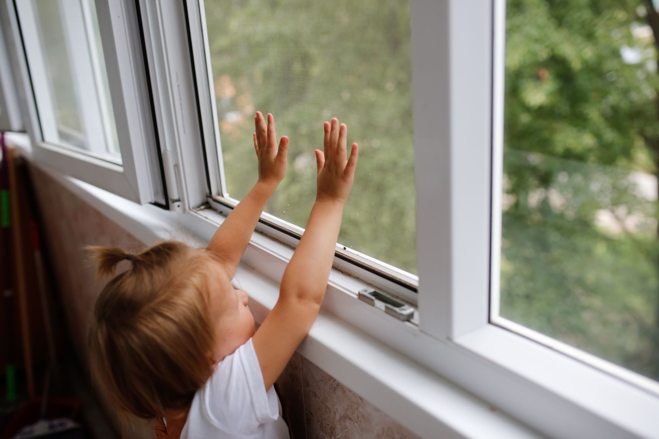 Kleinkind steht am Fenster und drückt mit den Händen gegen ein Fliegengitter.
