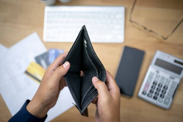 Zwei Hände halten eine leere Geldbörse über einem Schreibtisch mit Bankkarten und Taschenrechner.