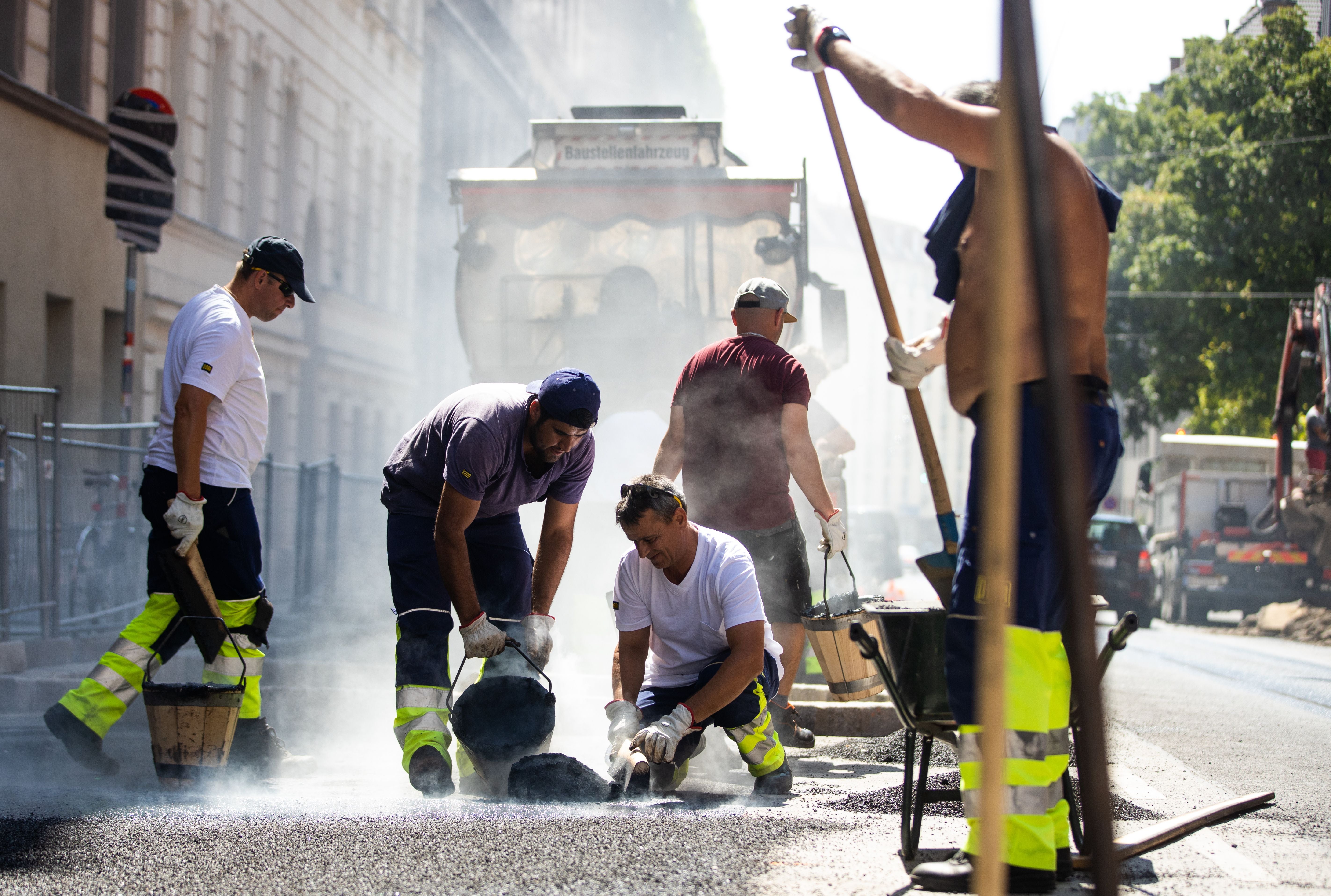 Straßenbauarbeiter arbeiten bei Hitze an einer Baustelle in Wien, umgeben von Staub und Maschinen.